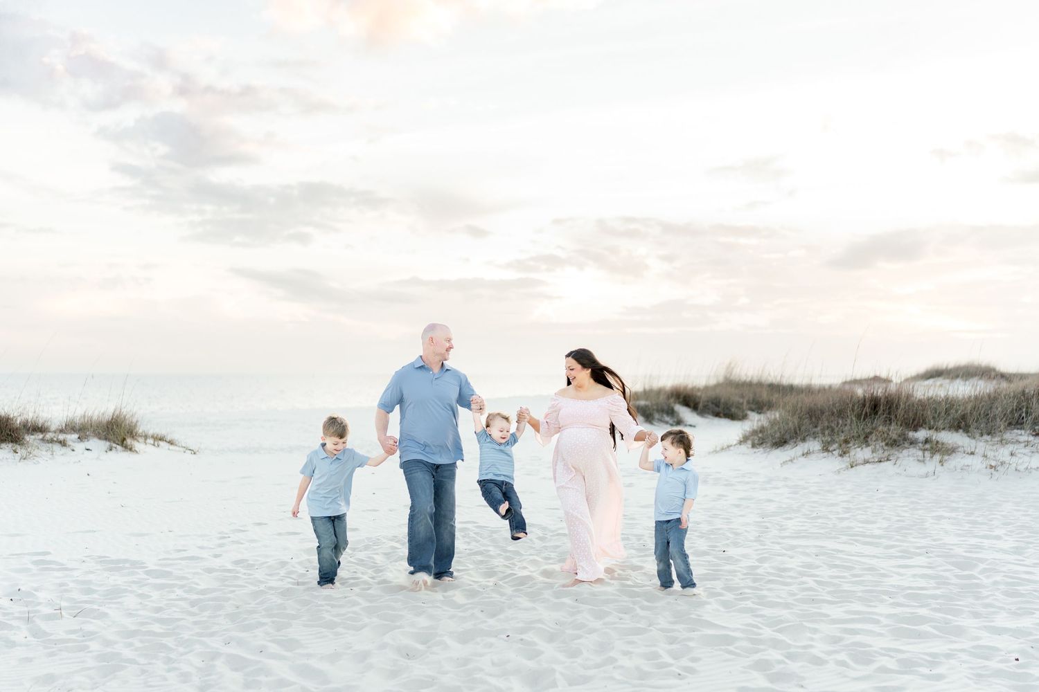 Family of five walks on white sand beach at sunset, parents holding children's hands, pregnant mother in flowing pink dress.