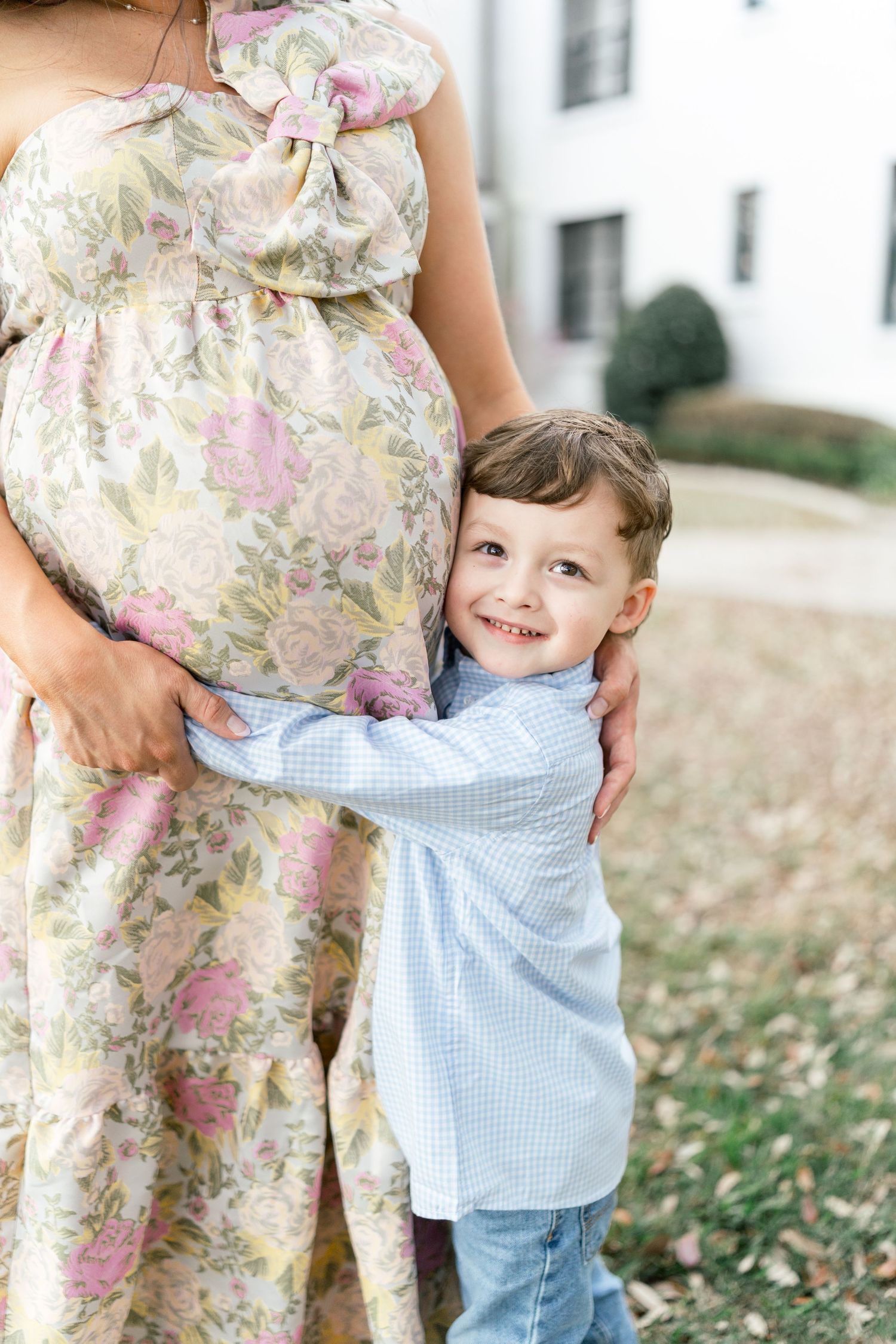A young boy smiles while hugging his pregnant mother's belly outdoors, wearing a blue shirt and jeans.