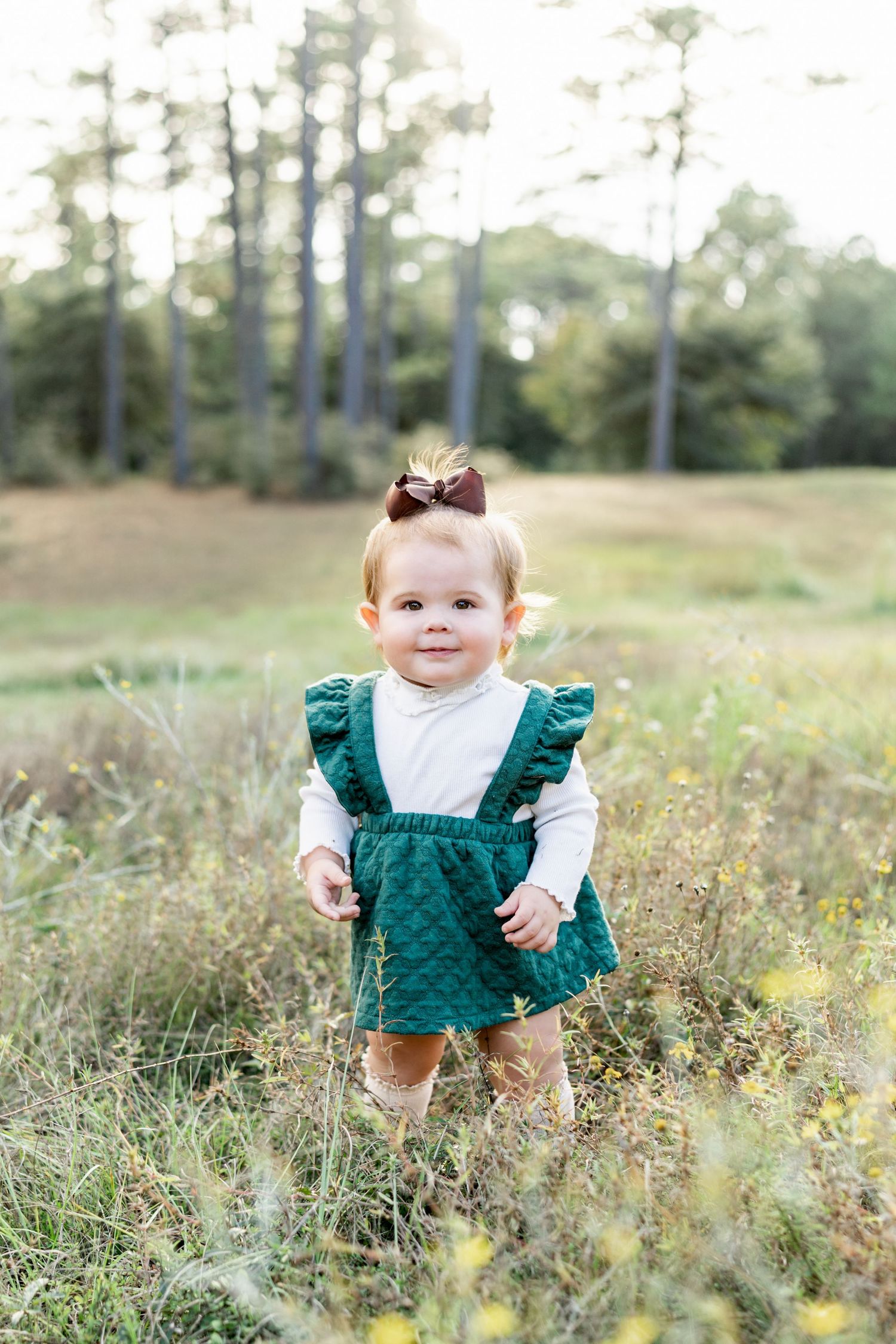 A small child in a green ruffled dress stands in a sunny meadow filled with wildflowers and tall grass located in Hattiesburg, Mississippi