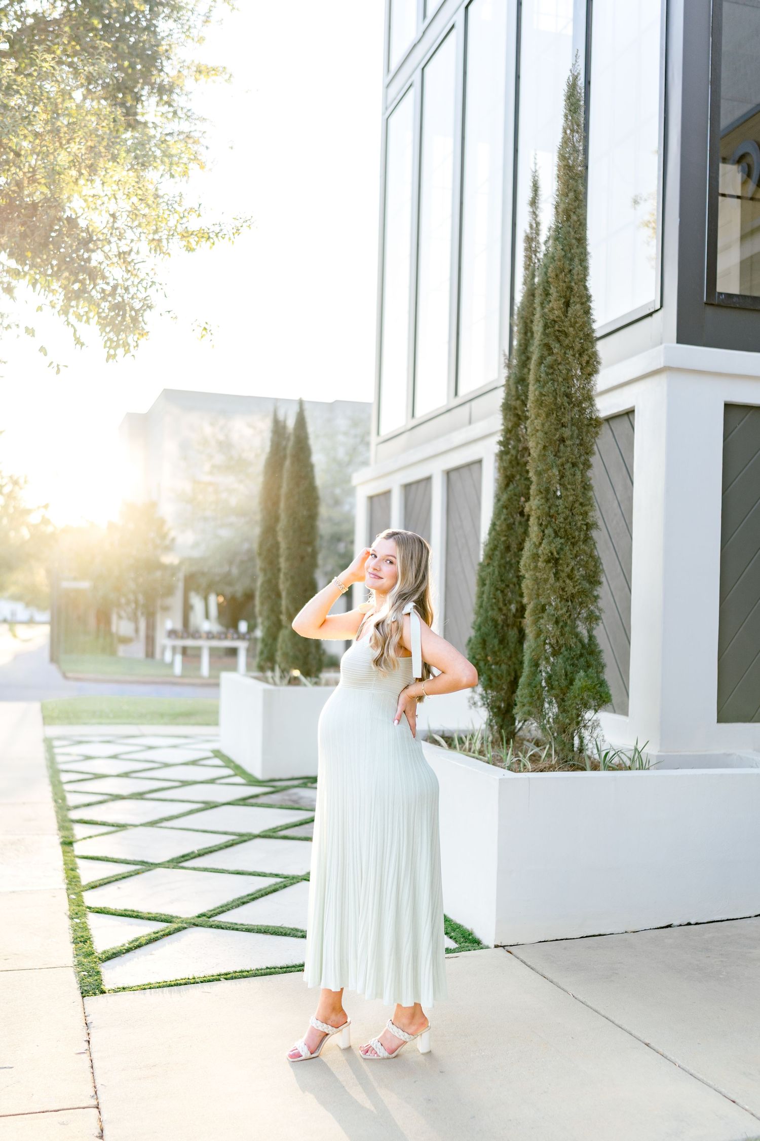 Woman in mint pleated maxi dress and white heels posing outside modern building with cypress trees in golden hour light.