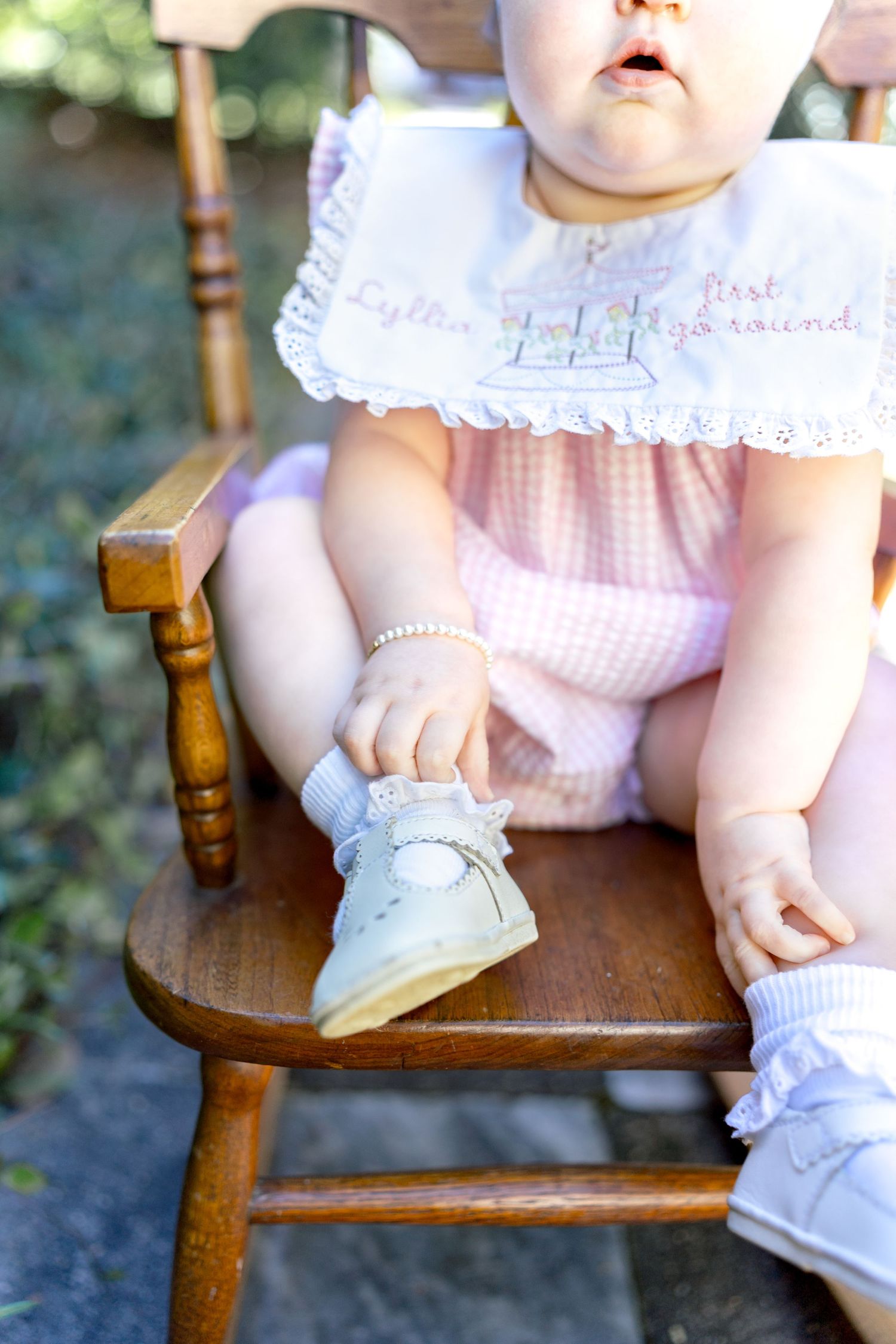 Baby girl in pink gingham outfit sitting in wooden chair, holding white shoe, wearing pearl bracelet.