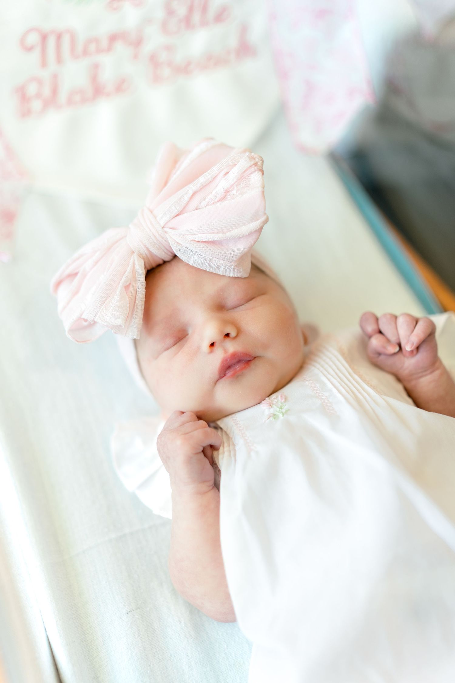 Sleeping newborn baby girl wearing a pink bow headband, dressed in white, lying in a crib with a personalized blanket.
