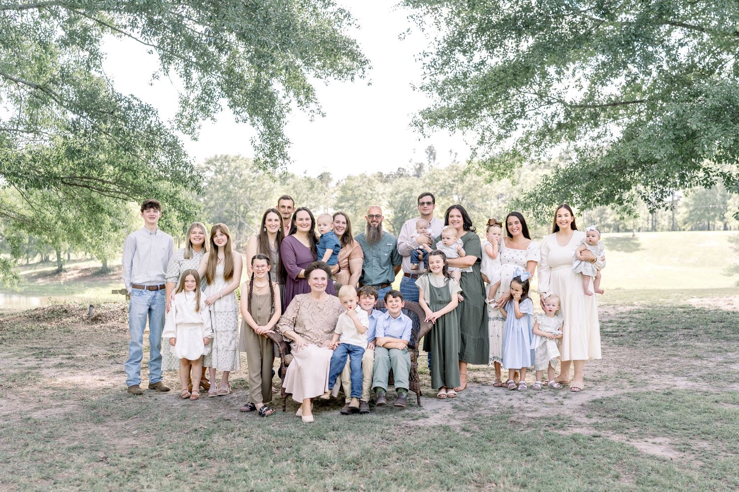 Large extended family group portrait outdoors under trees in a park, wearing coordinated neutral outfits.