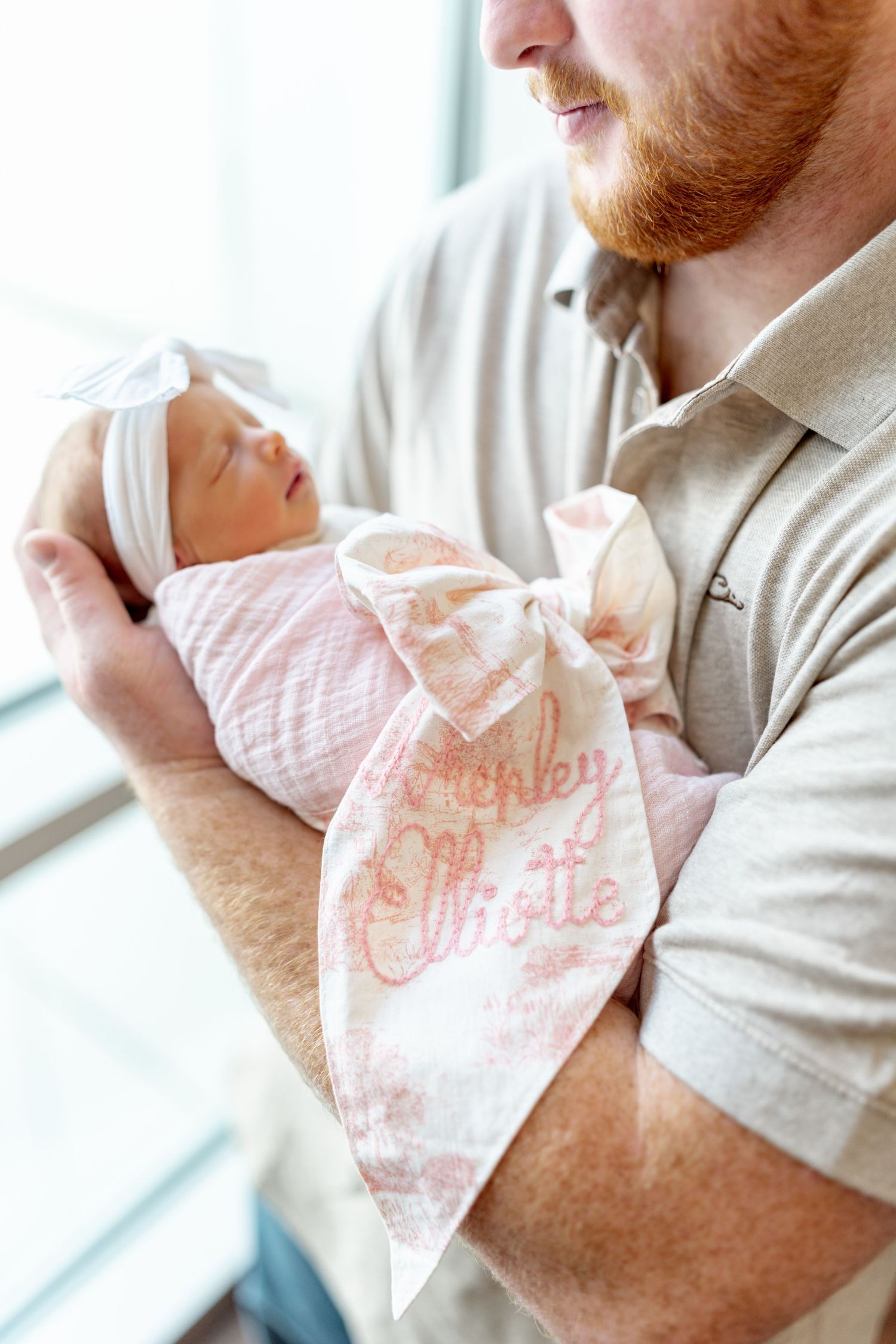 A red-bearded father cradles his newborn baby girl wrapped in a personalized pink swaddle blanket reading Ashley Charlotte.