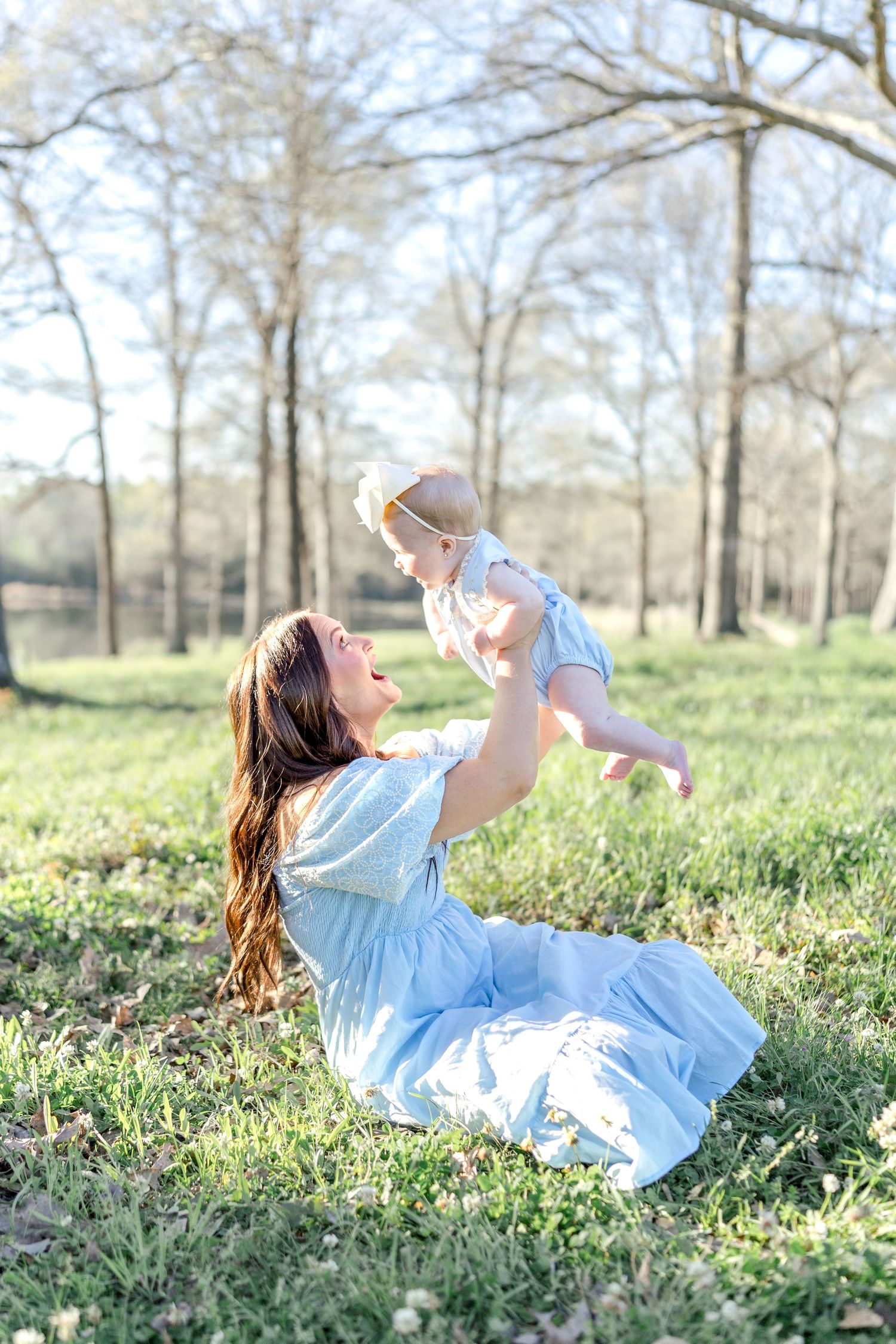 A mother in a blue dress sits on grass, joyfully lifting her baby in the air outdoors.