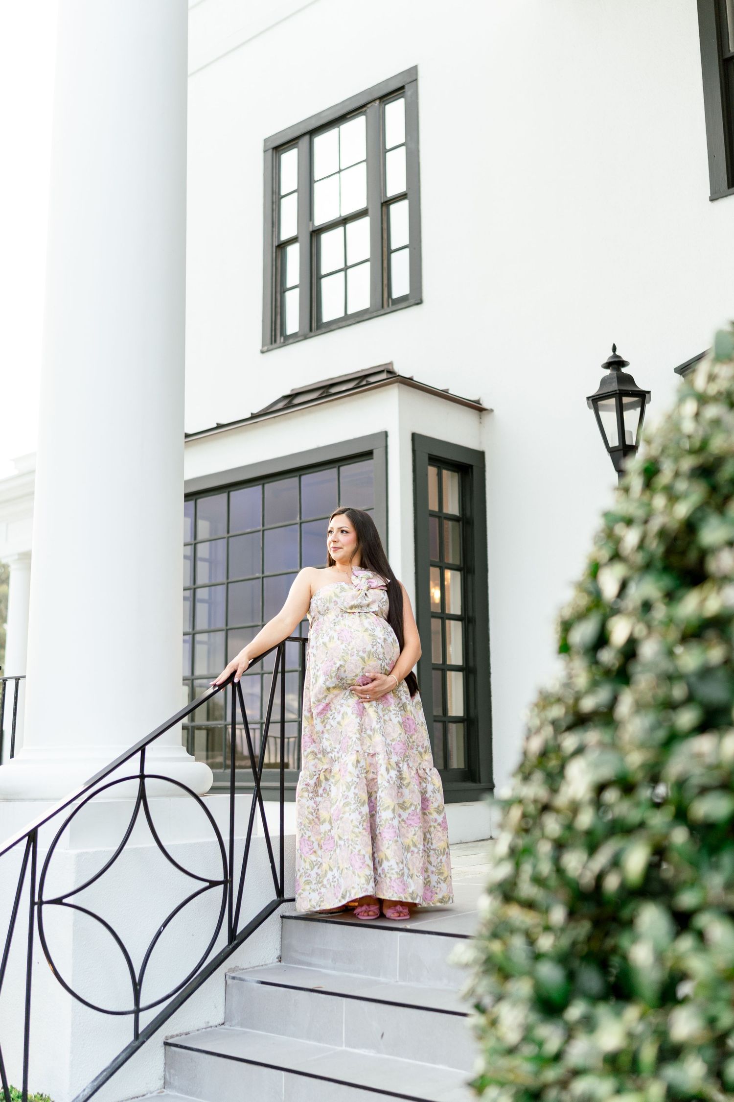 Pregnant woman in floral maxi dress standing on white mansion steps, cradling baby bump, elegant outdoor maternity photo.