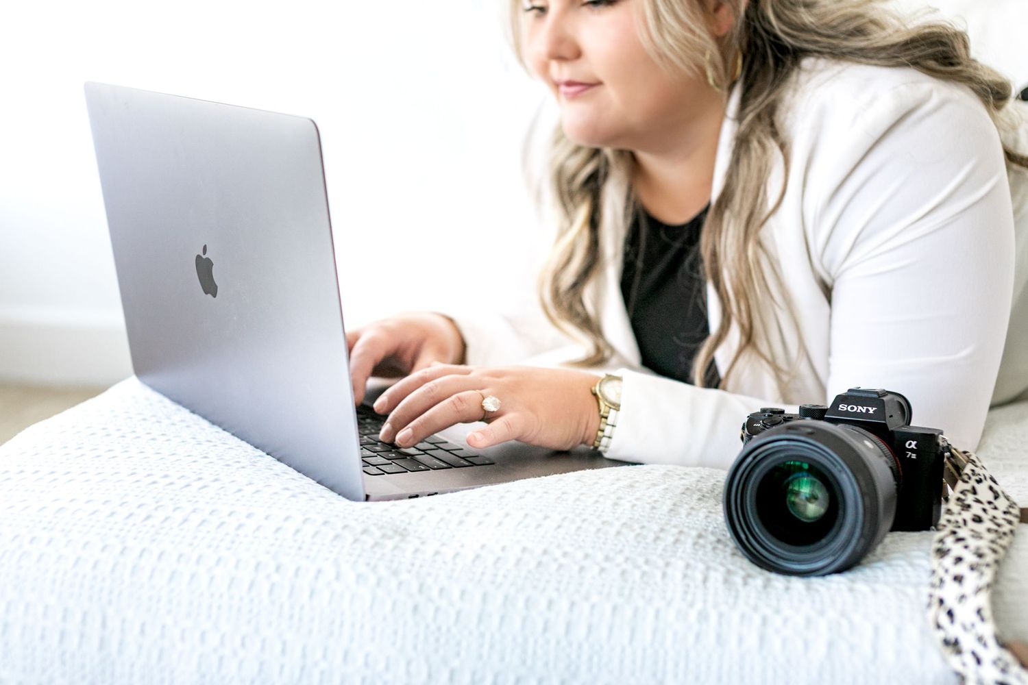 Photographer working on laptop while lying on white bedding with camera nearby.