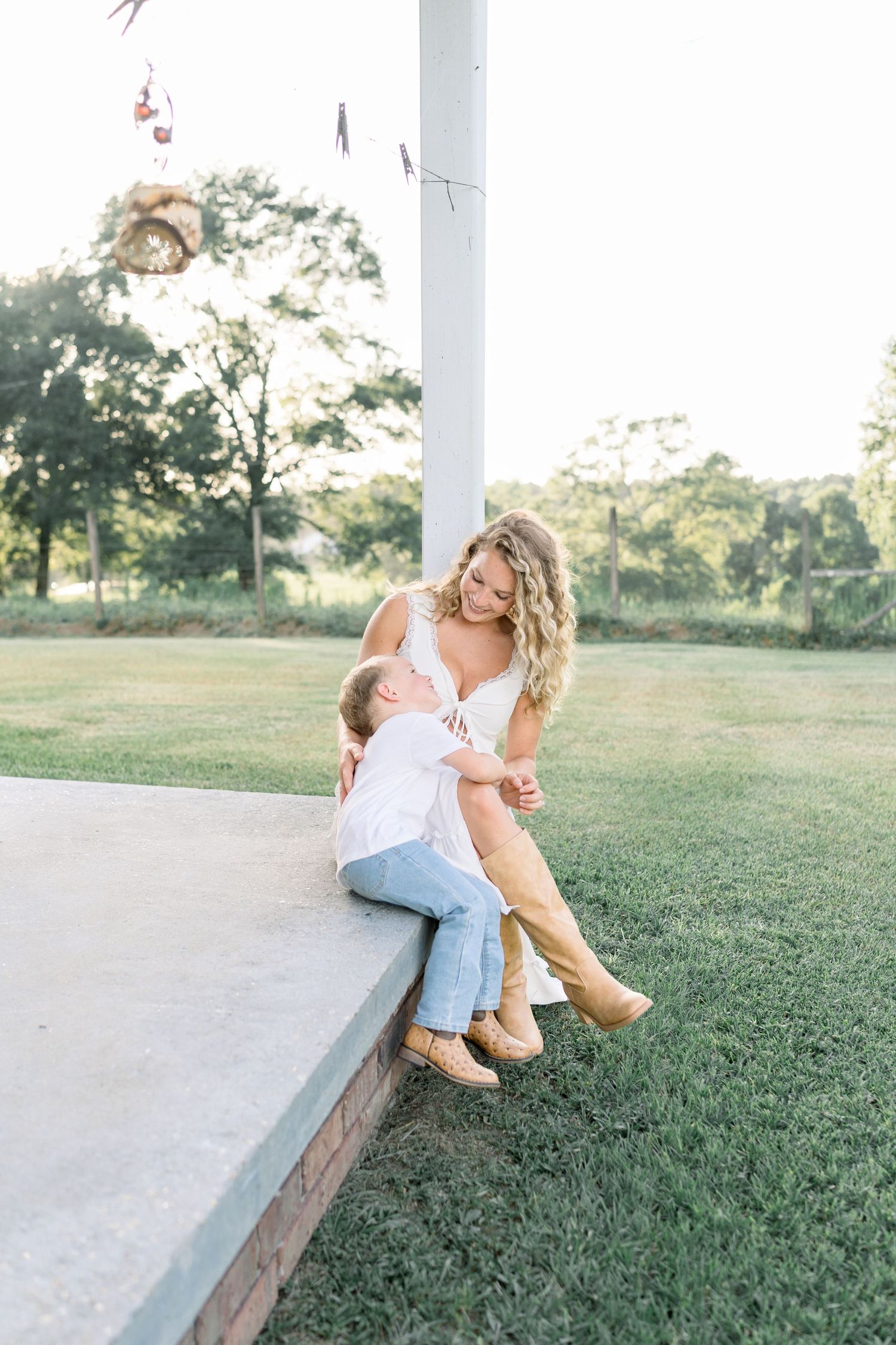 A woman with curly blonde hair sits on a porch ledge with a young child in a sunny green yard.