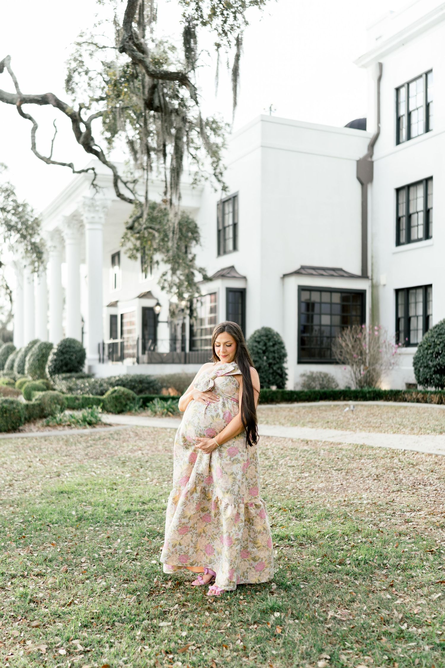 Pregnant woman in floral maxi dress cradling baby bump outdoors in front of elegant white Southern mansion.