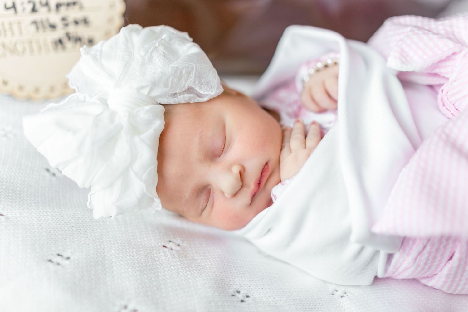 Peaceful newborn baby sleeping swaddled in white blanket wearing a white bow headband and pink striped outfit.