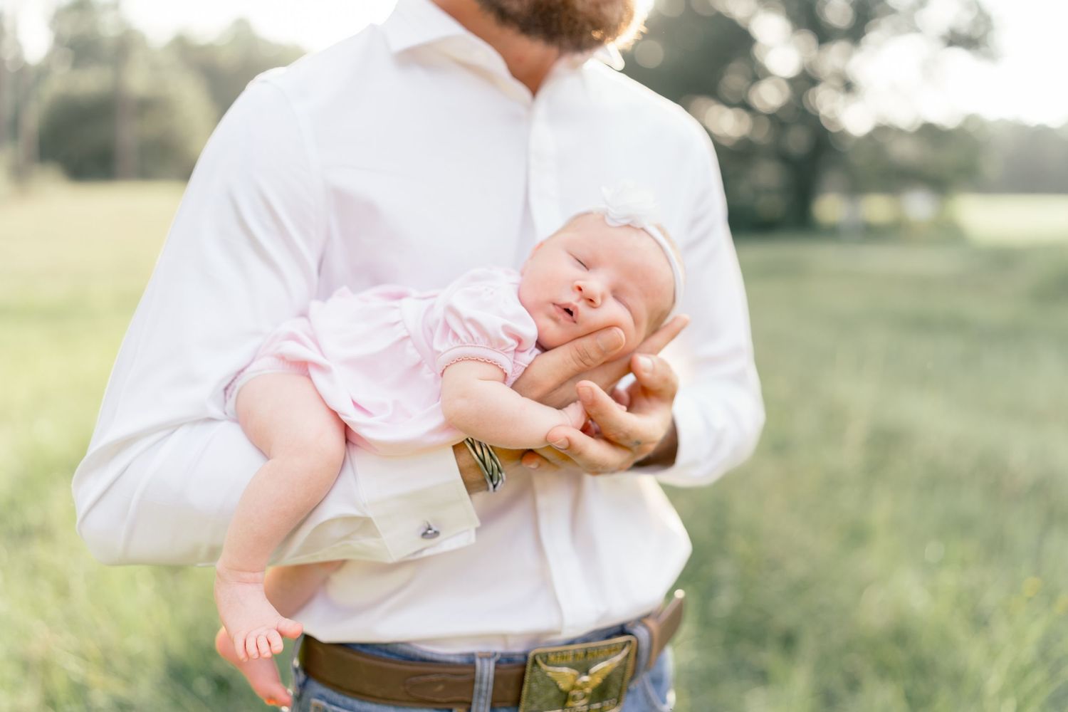 Father in white shirt cradling sleeping newborn baby in pink outfit outdoors in green field during golden hour.
