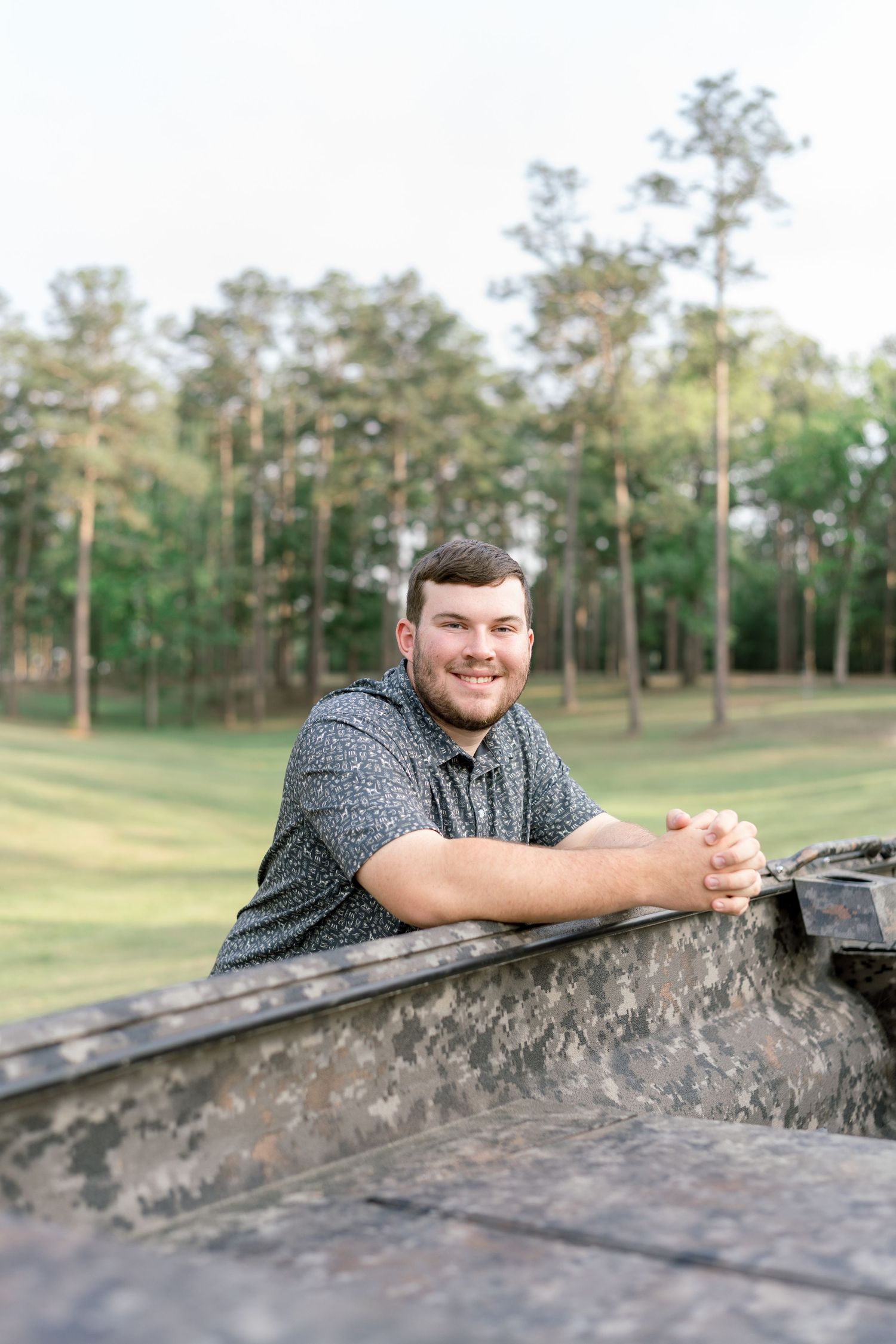 Young man smiling while leaning on a camouflage truck bed outdoors with tall pine trees in the background.