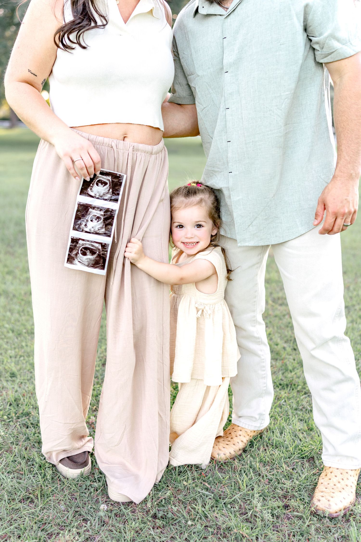 Pregnant woman holding ultrasound photos with toddler girl hugging her leg beside father outdoors.