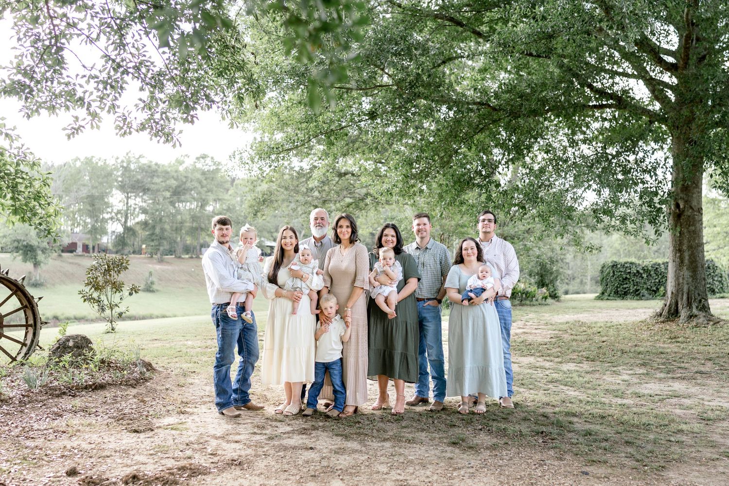 Large family group portrait outdoors under oak trees on a green lawn, dressed in coordinated neutral tones.