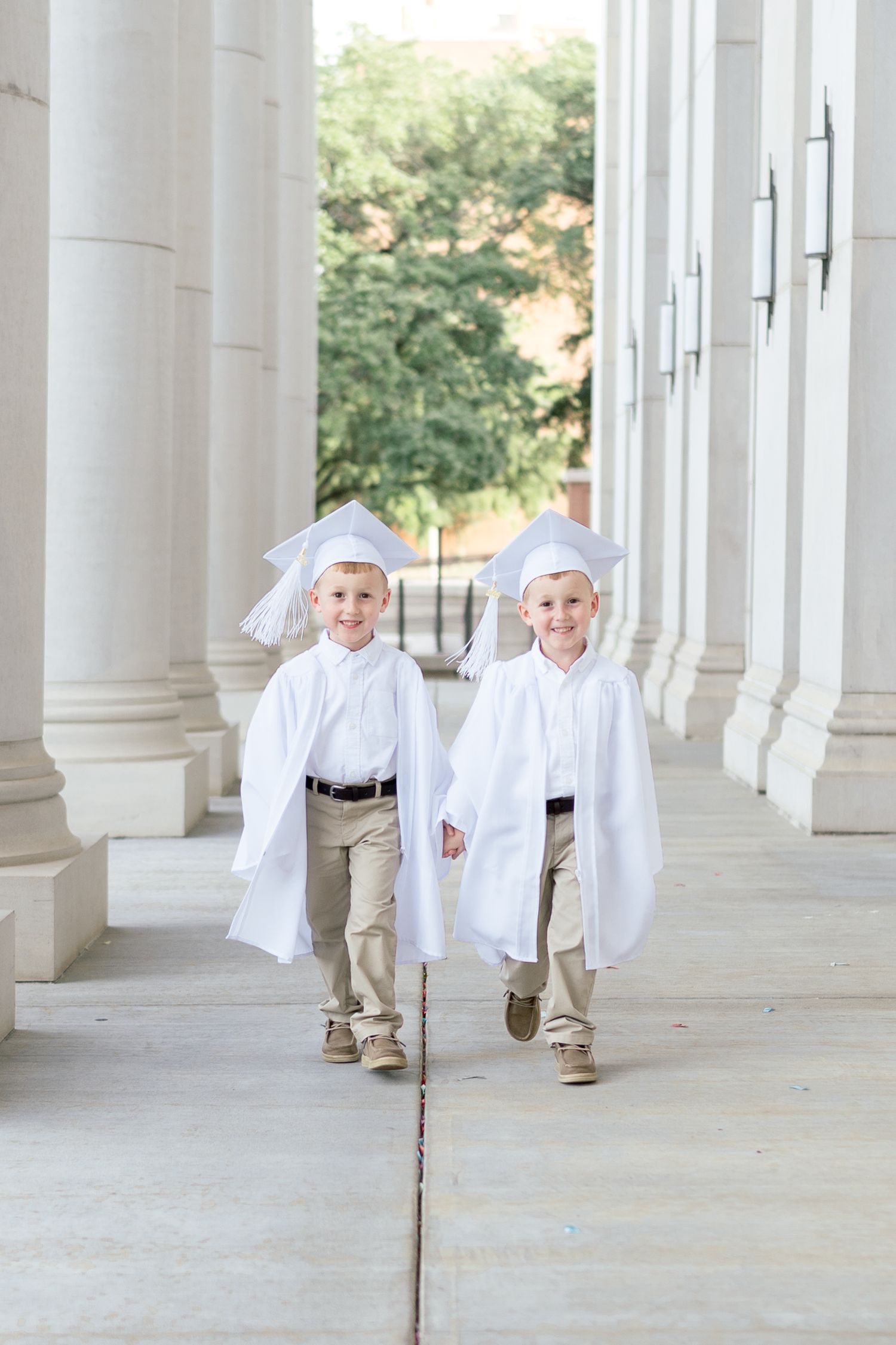 Two young boys in white graduation caps and gowns walking between white columns at a graduation ceremony.