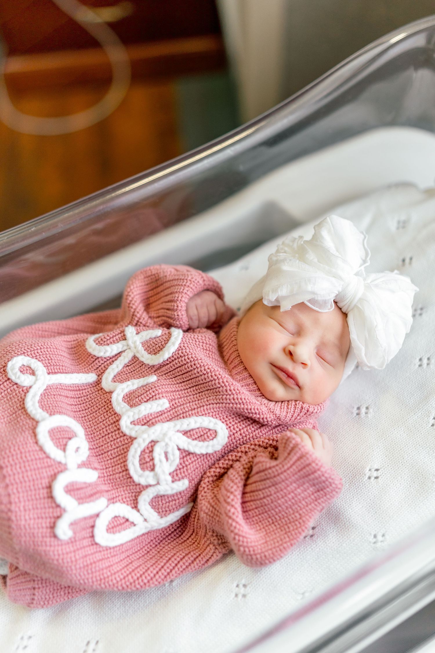 Newborn baby girl sleeping in hospital bassinet wearing pink knit romper and white bow headband.