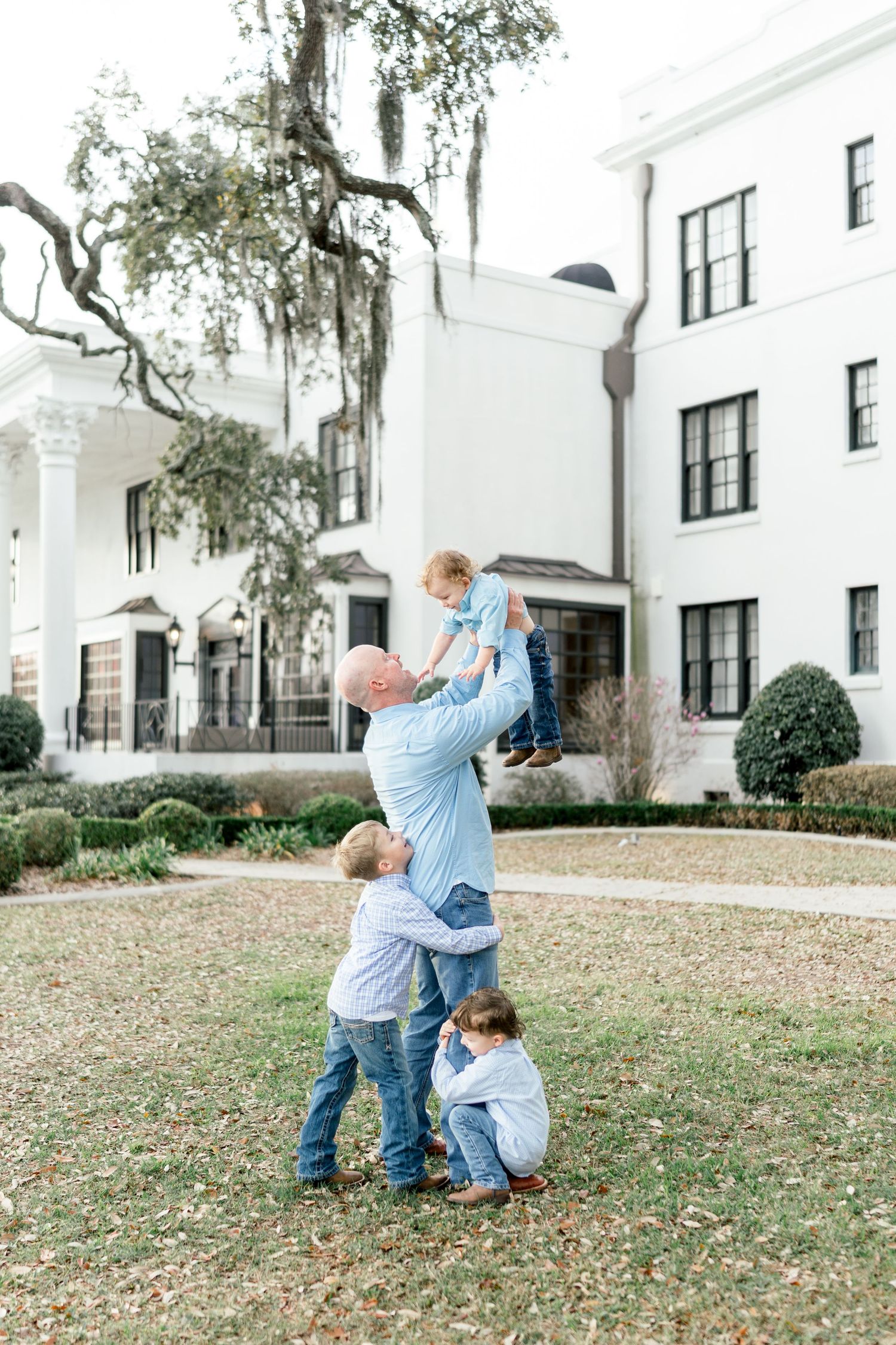 A father lifts a toddler in the air while two children play around him outside a white Southern mansion.