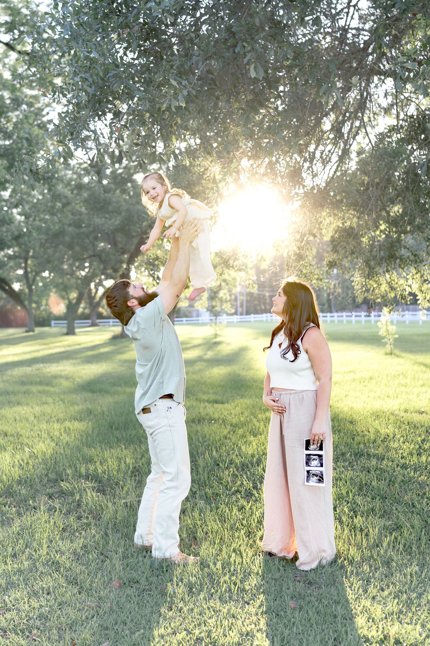 A man lifts a toddler in the air while a pregnant woman holds ultrasound photos in a sunny park setting.