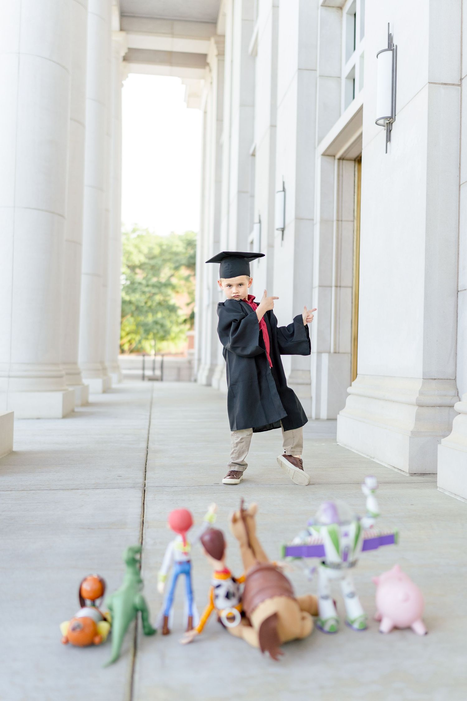 Young child in graduation cap and gown poses proudly outdoors with Toy Story figurines arranged in foreground.
