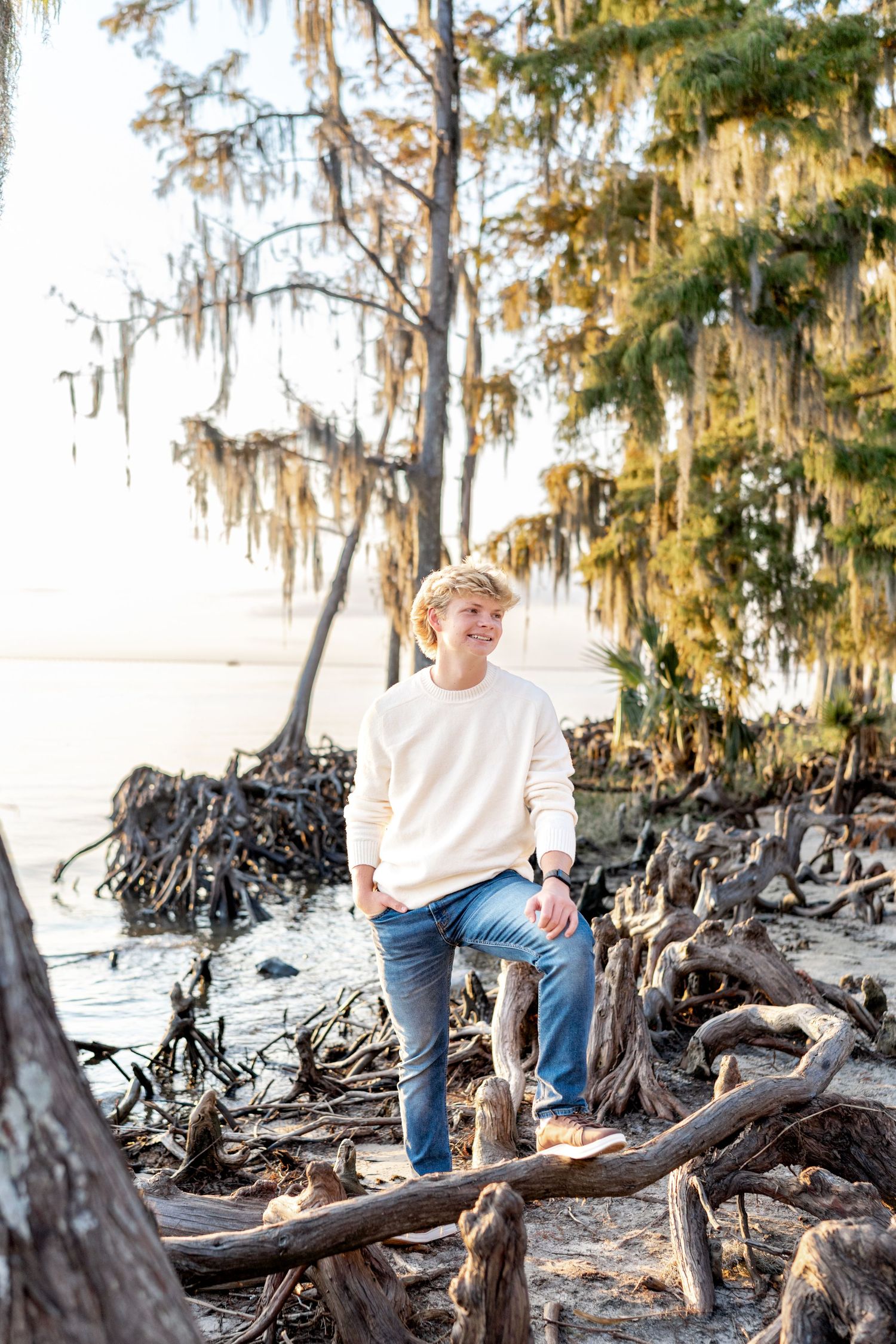 Person in a cream sweater and jeans sits on driftwood along a scenic beach with Spanish moss-draped trees.