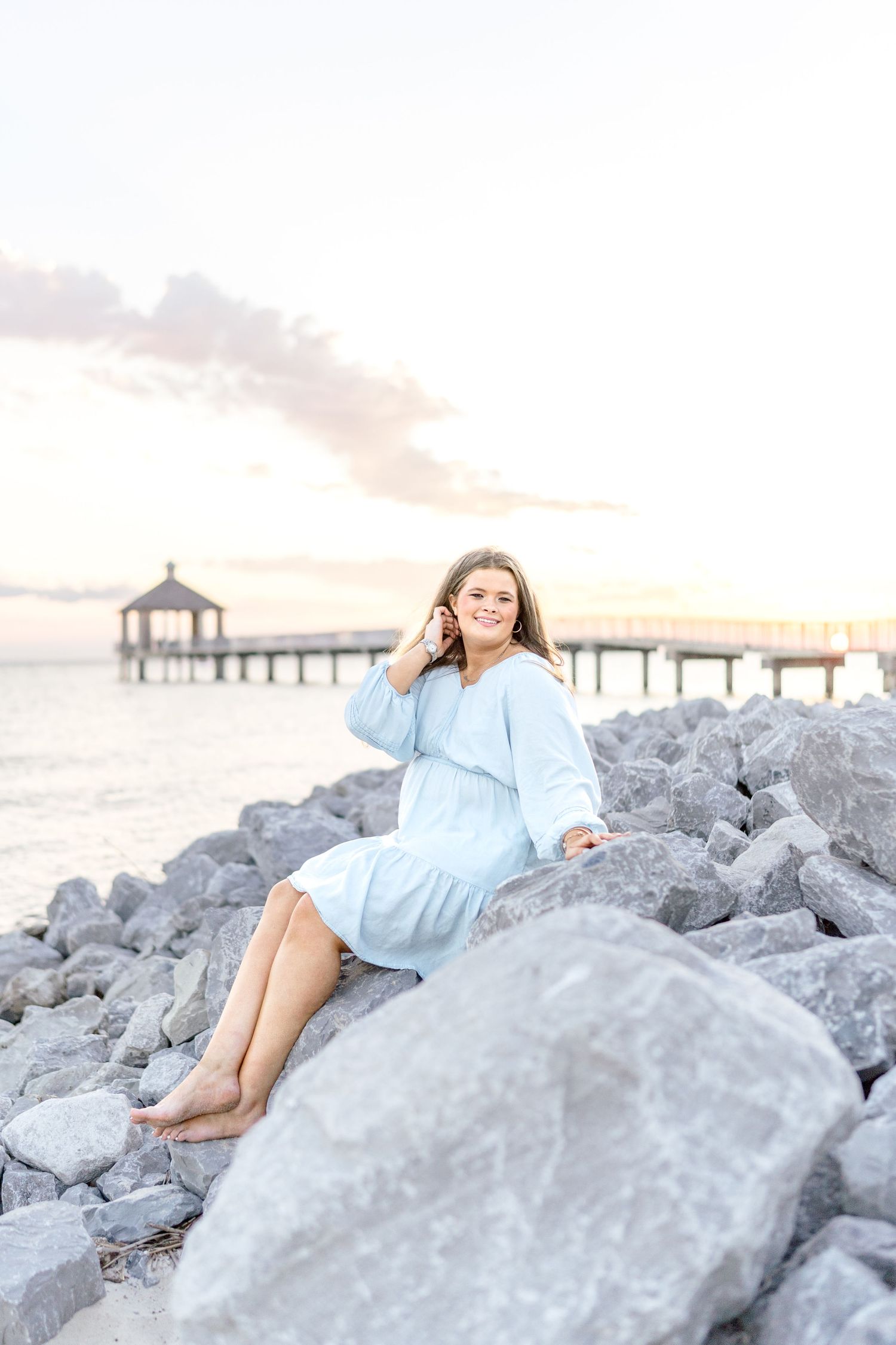 Young woman in light blue dress sitting on rocky shoreline at sunset with pier in background.