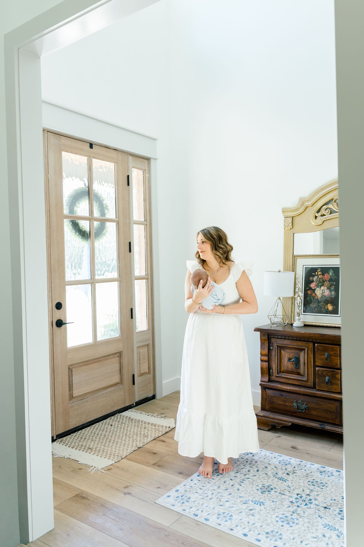 A woman in a white dress holds a newborn baby near a wooden front door with a wreath in a bright, elegant home.