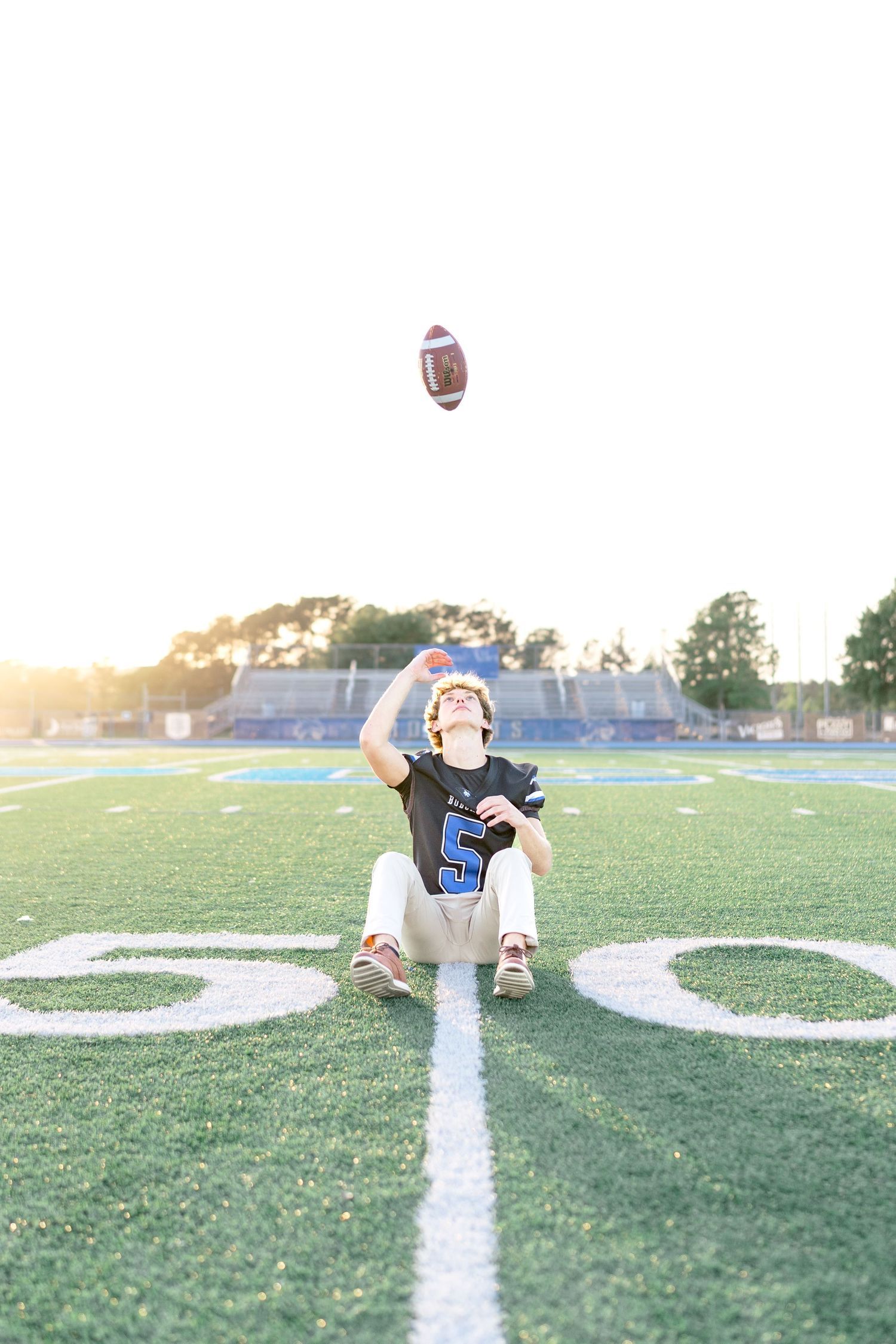 Football player #5 sits on the 50-yard line tossing a football in the air at sunset on a green turf field.