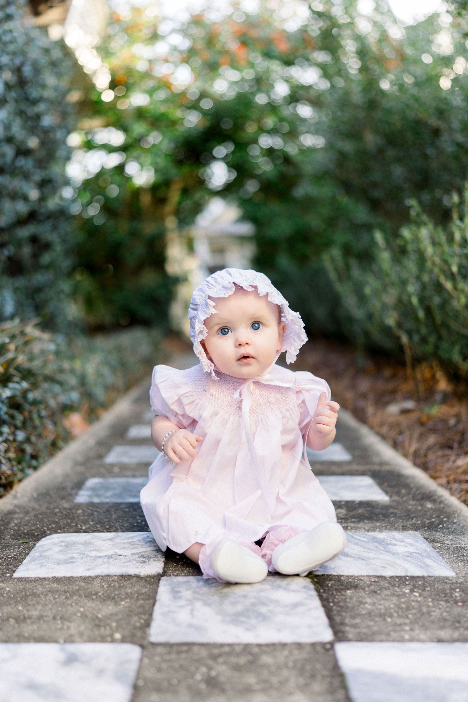 Adorable baby girl in pink dress and bonnet sitting on checkered stone path surrounded by lush green garden.