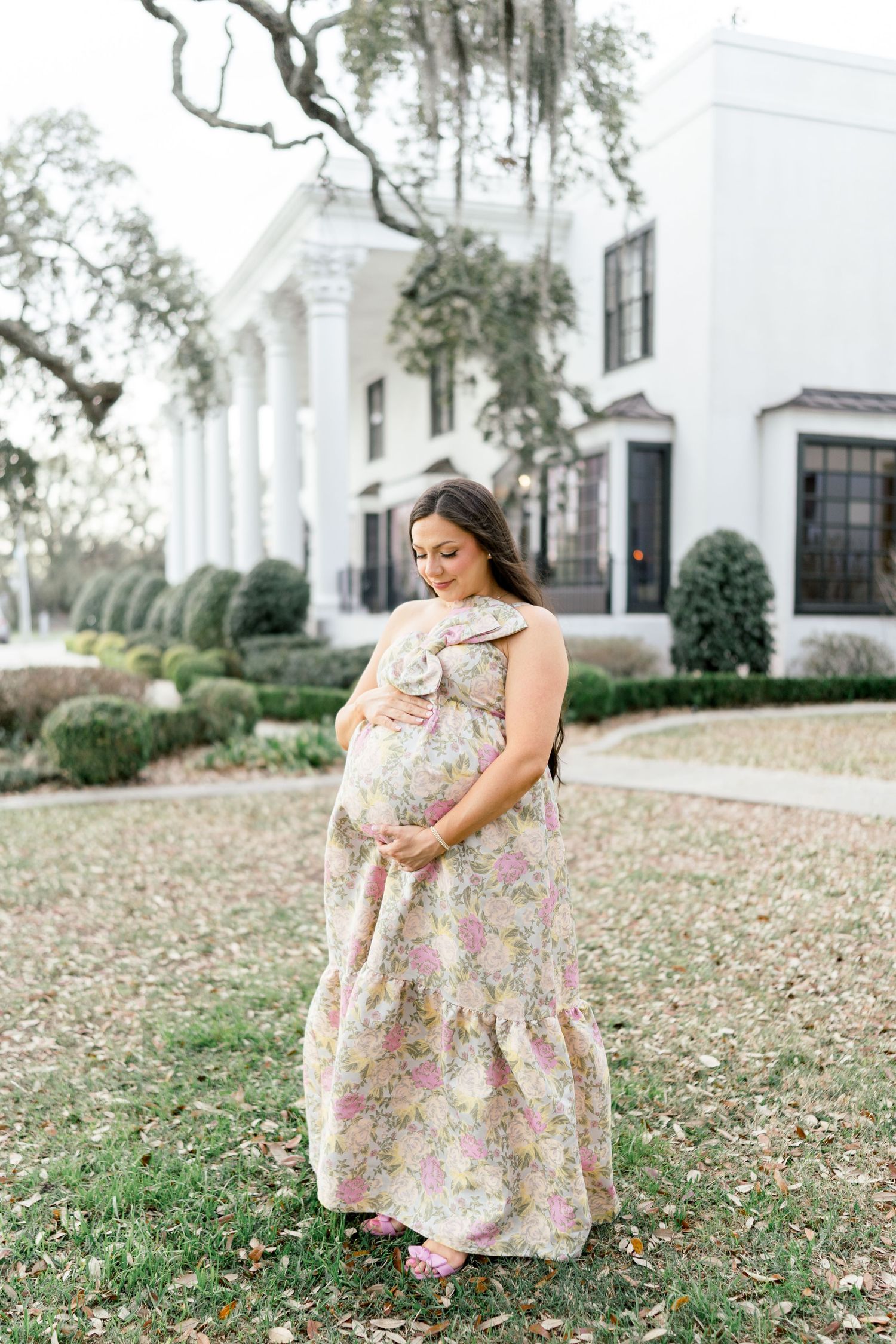 Pregnant woman in floral maxi dress cradling baby bump outdoors in front of elegant white Southern mansion.