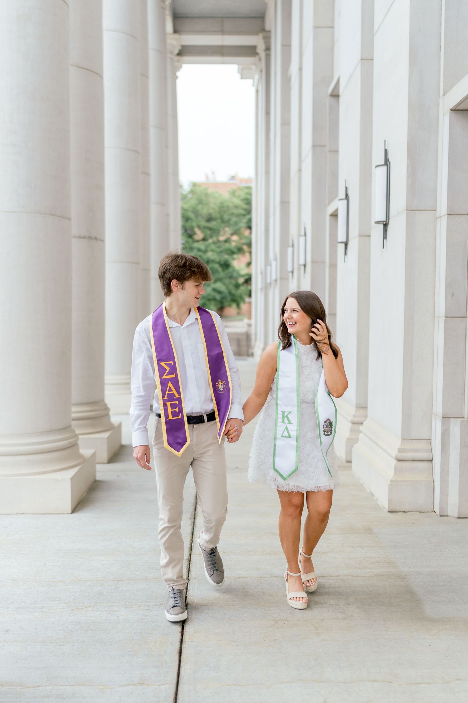 Two college graduates in Greek stoles walk hand in hand between white columns, smiling at each other.