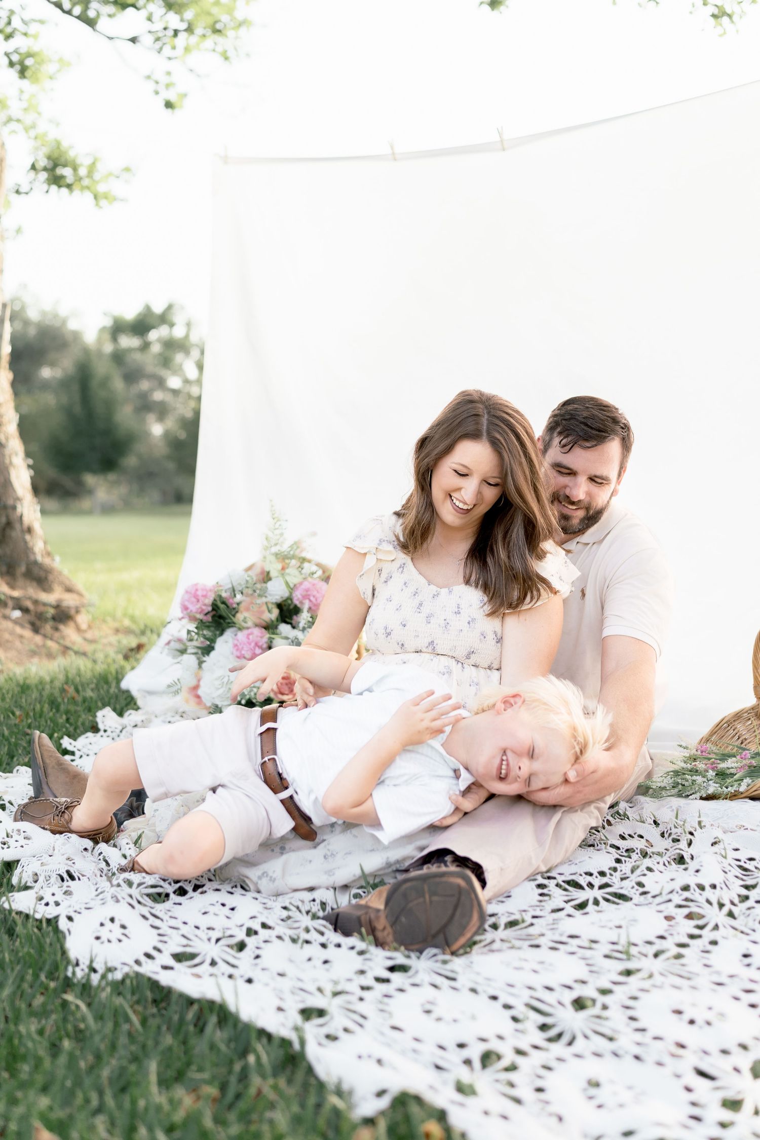 A joyful family of three laughs together on a lace blanket outdoors, with a white backdrop and pink flowers nearby.