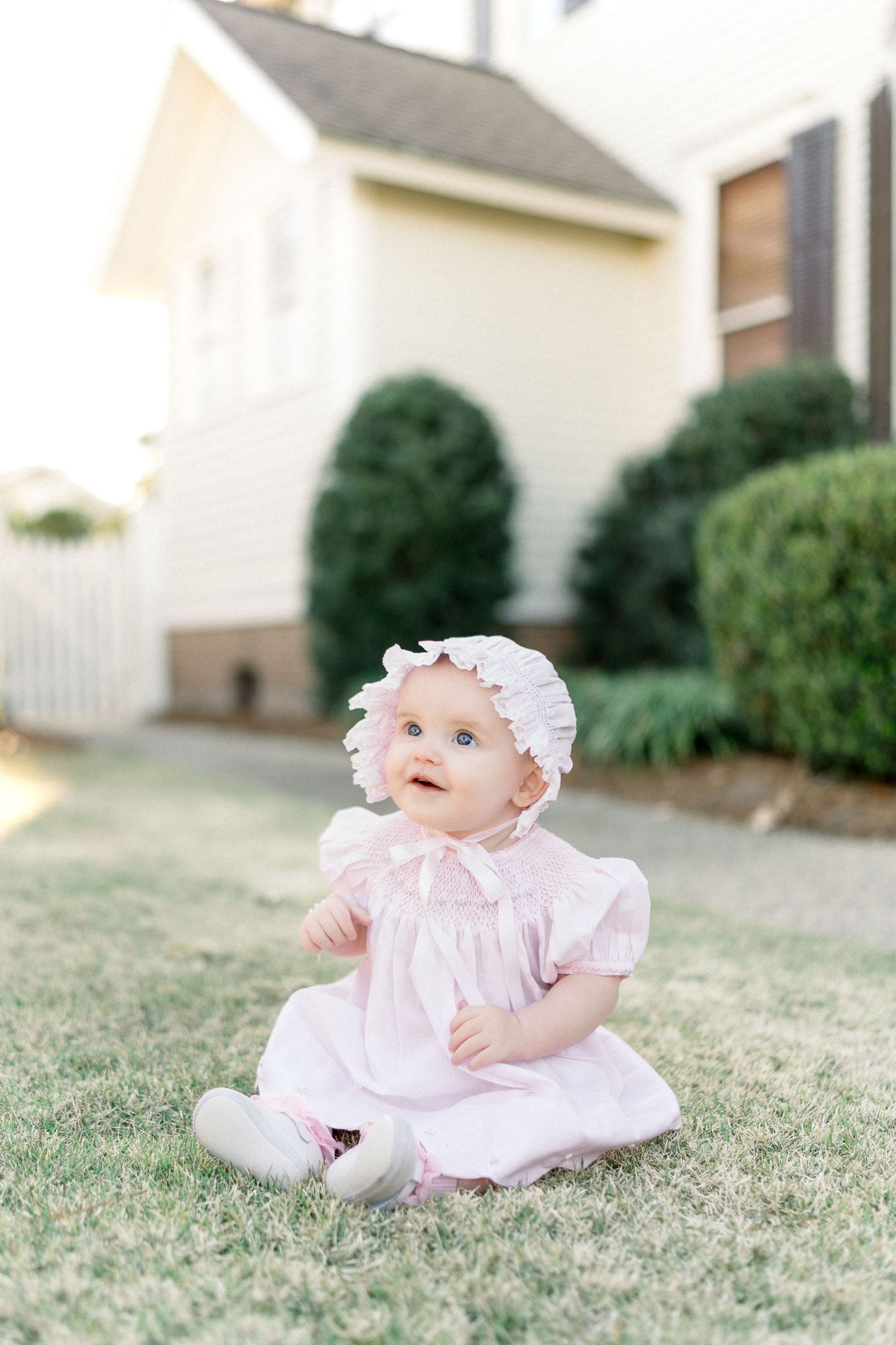 Adorable baby girl in pink dress and bonnet sitting on green grass outdoors near a white house.