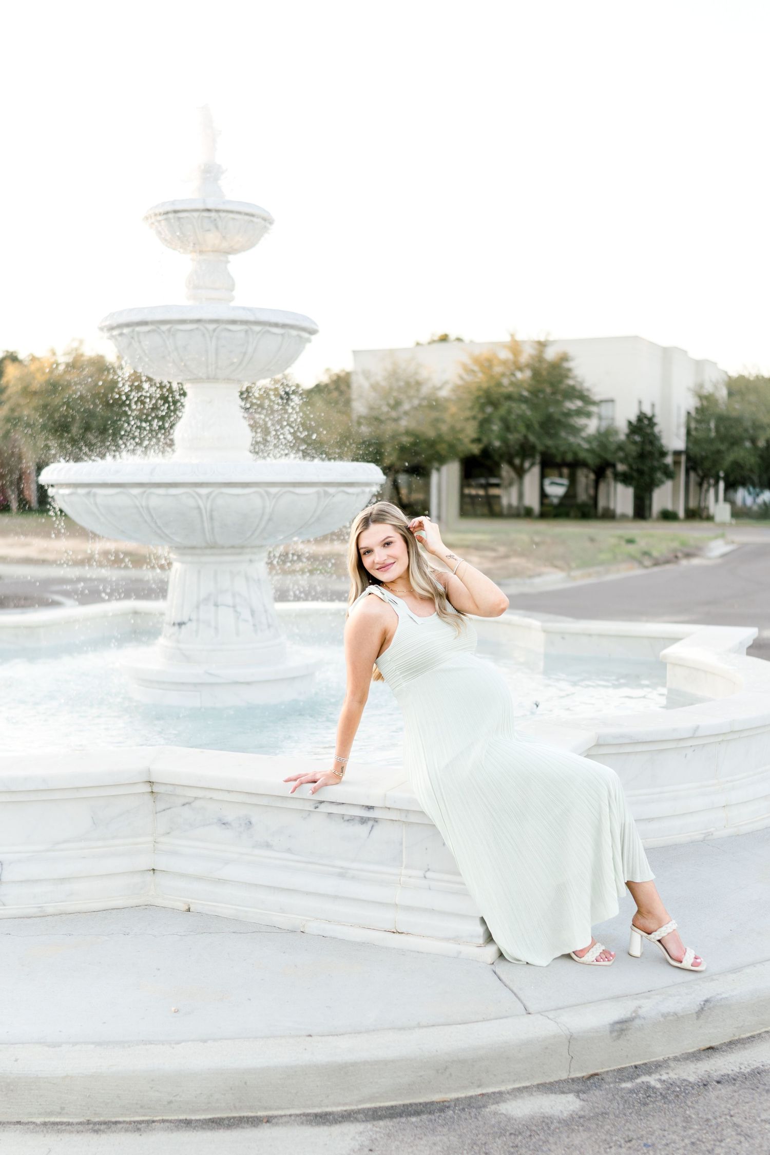 Young woman in mint green dress poses elegantly by a white tiered fountain outdoors.