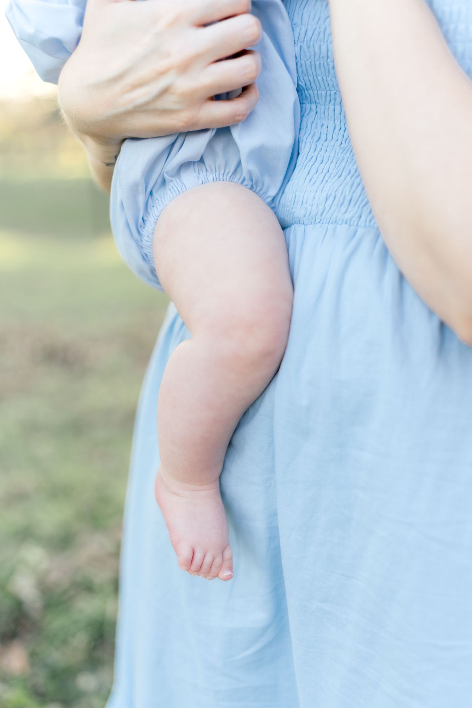 Close-up of a baby's chubby legs dangling while being held by a mother in a light blue smocked dress outdoors.