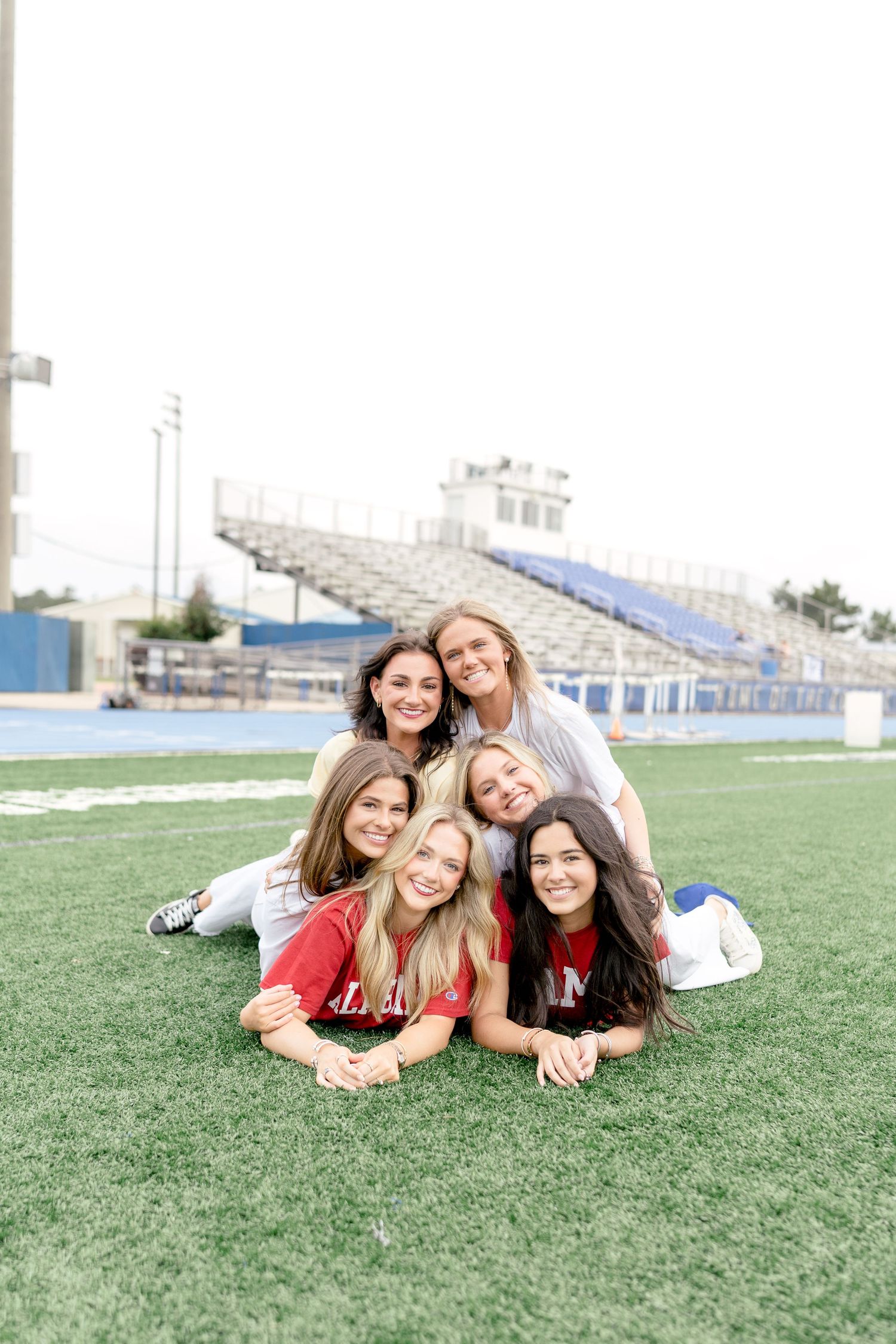 Six young women pose in a pyramid formation on a football field turf, smiling at the camera with stadium bleachers behind them.