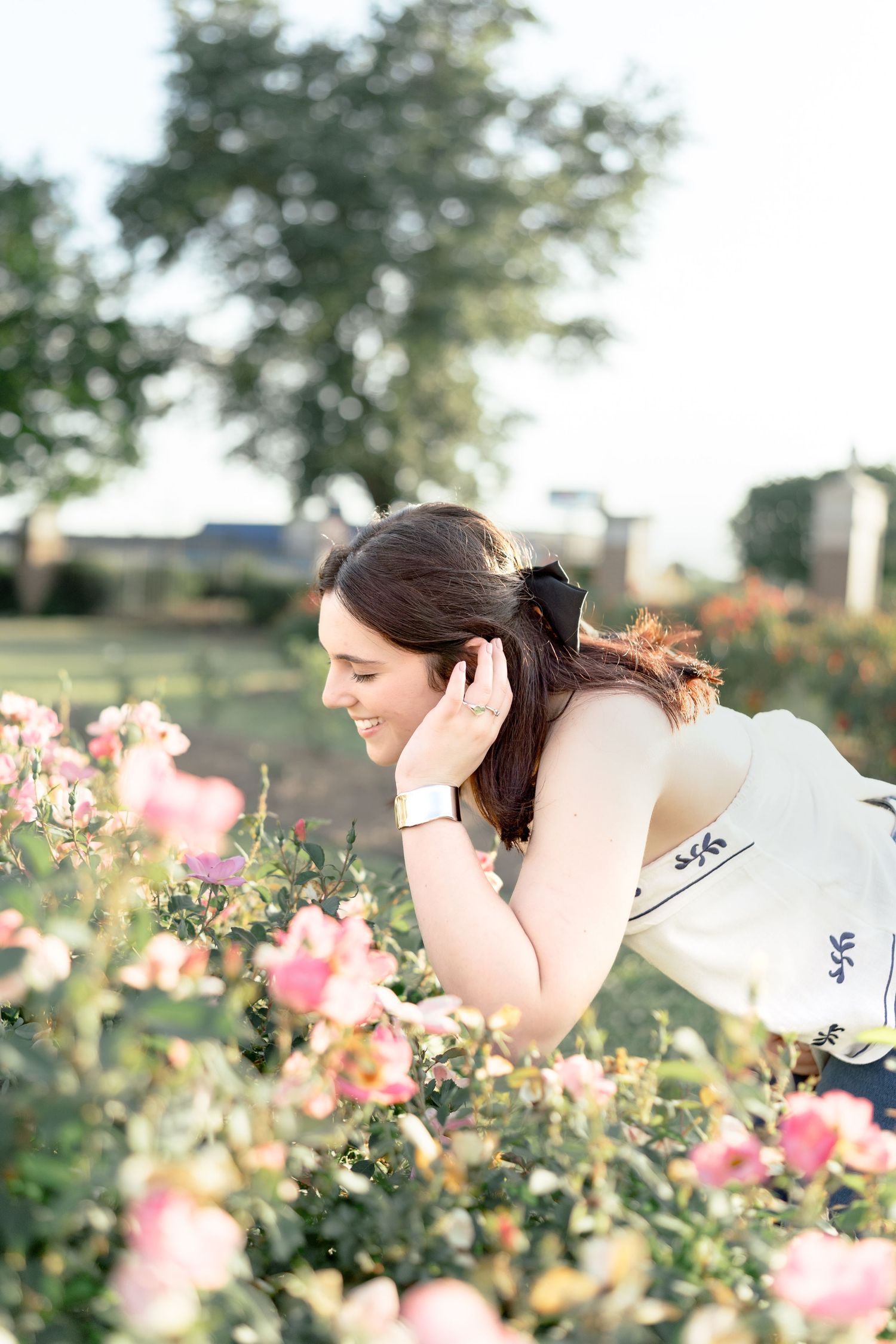 A smiling young woman in a white dress leans over blooming pink roses in a lush garden on a sunny day.