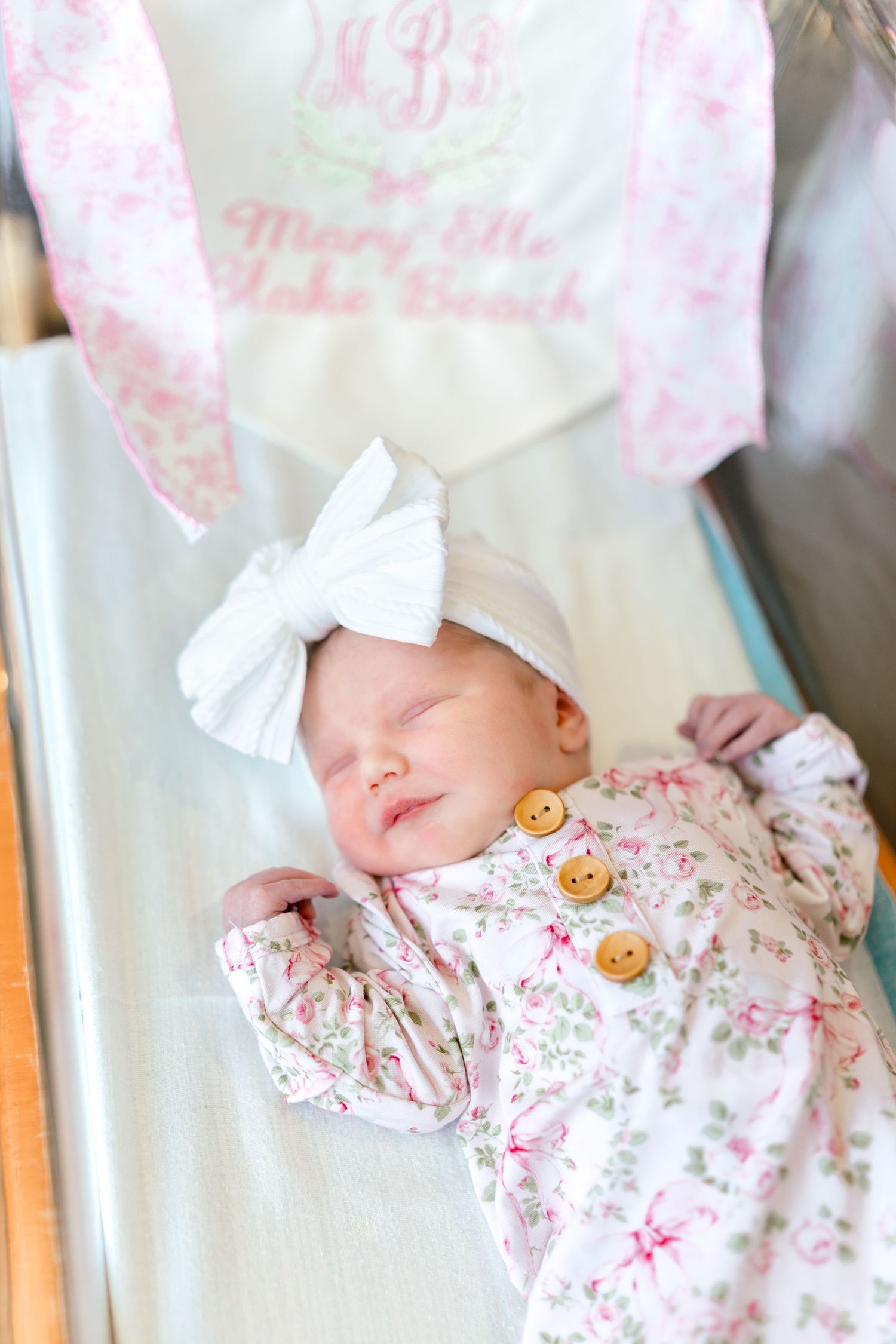 Newborn baby girl sleeping in hospital bassinet wearing floral outfit and white bow headband.