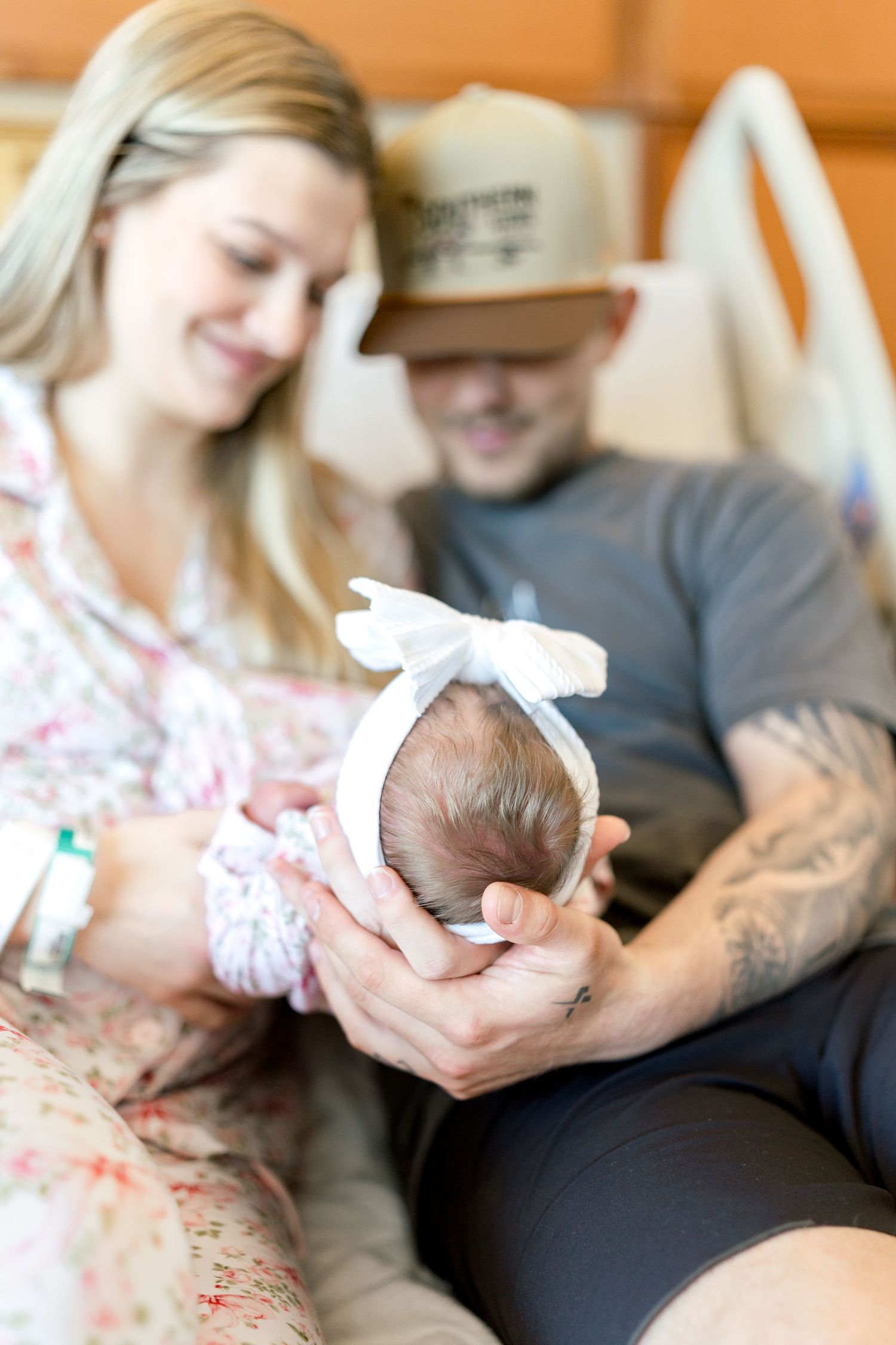 New parents tenderly hold their swaddled newborn baby girl wearing a white bow headband in a hospital room.