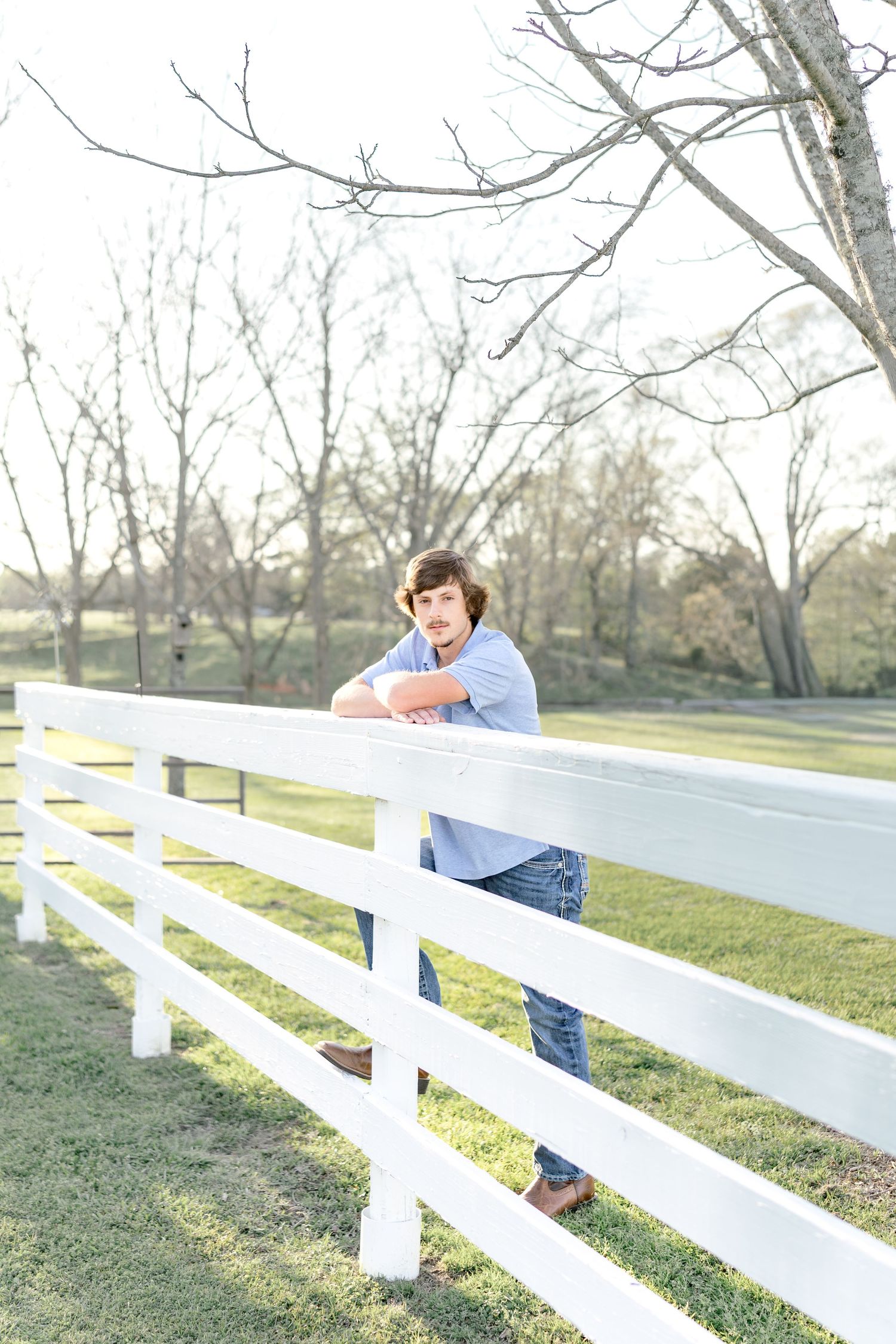 A young man in a blue shirt and jeans leans on a white wooden fence outdoors with bare trees in the background.