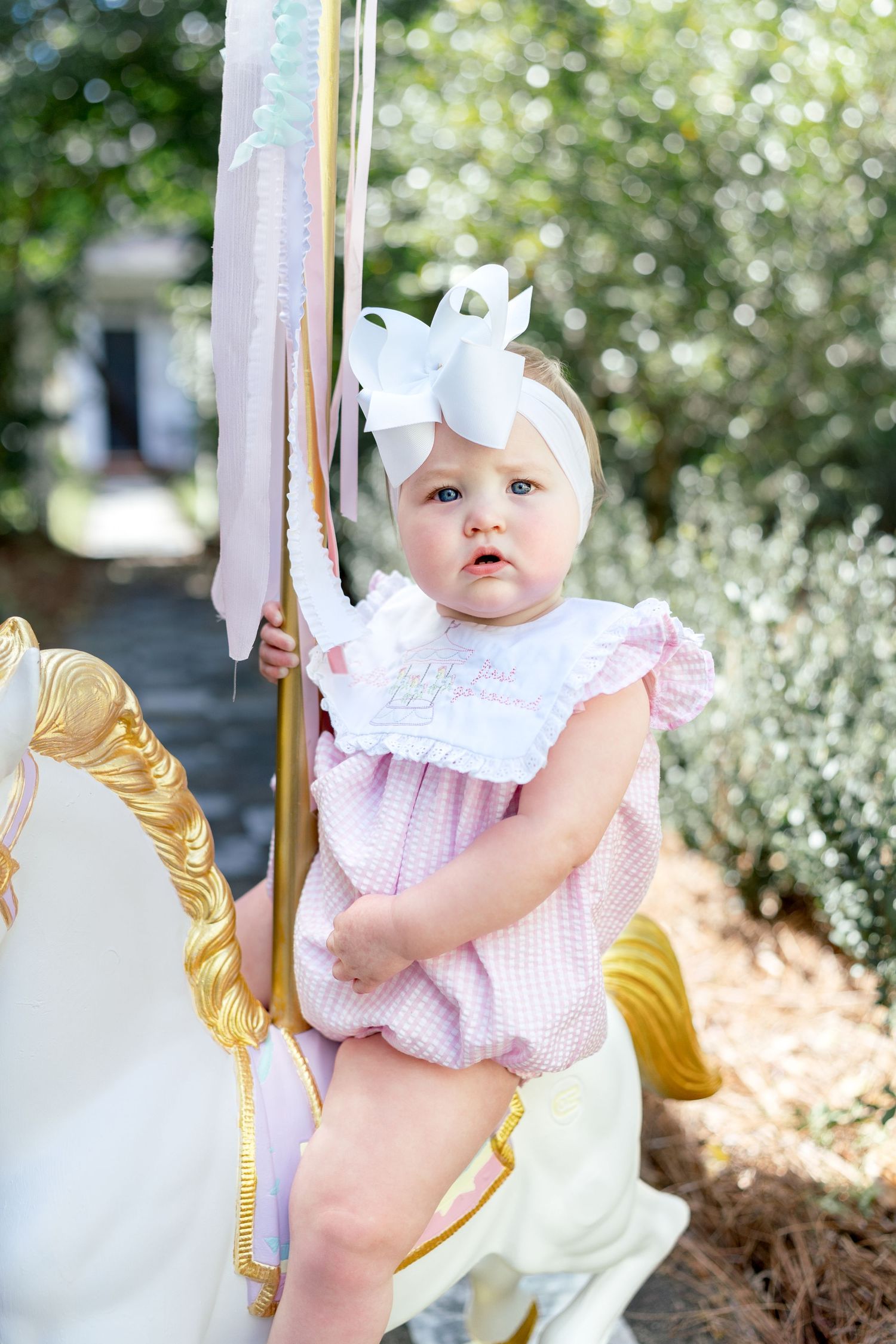 Baby girl in pink gingham romper with white bow headband sitting on a carousel horse outdoors.
