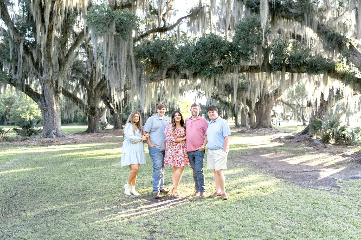 Family of five posing under sprawling oak trees draped with Spanish moss in a lush green park.