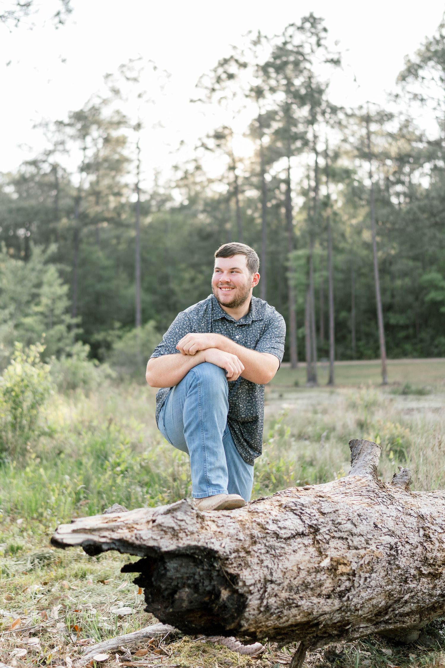 Young man in blue shirt and jeans smiling while perched on a fallen log in a forest clearing.