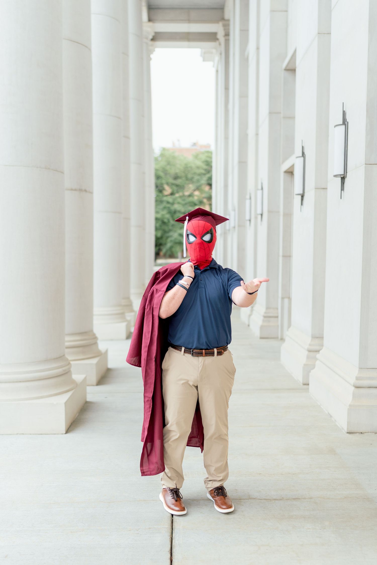Person wearing Spider-Man mask and graduation cap holds maroon robe beside white columns outdoors.