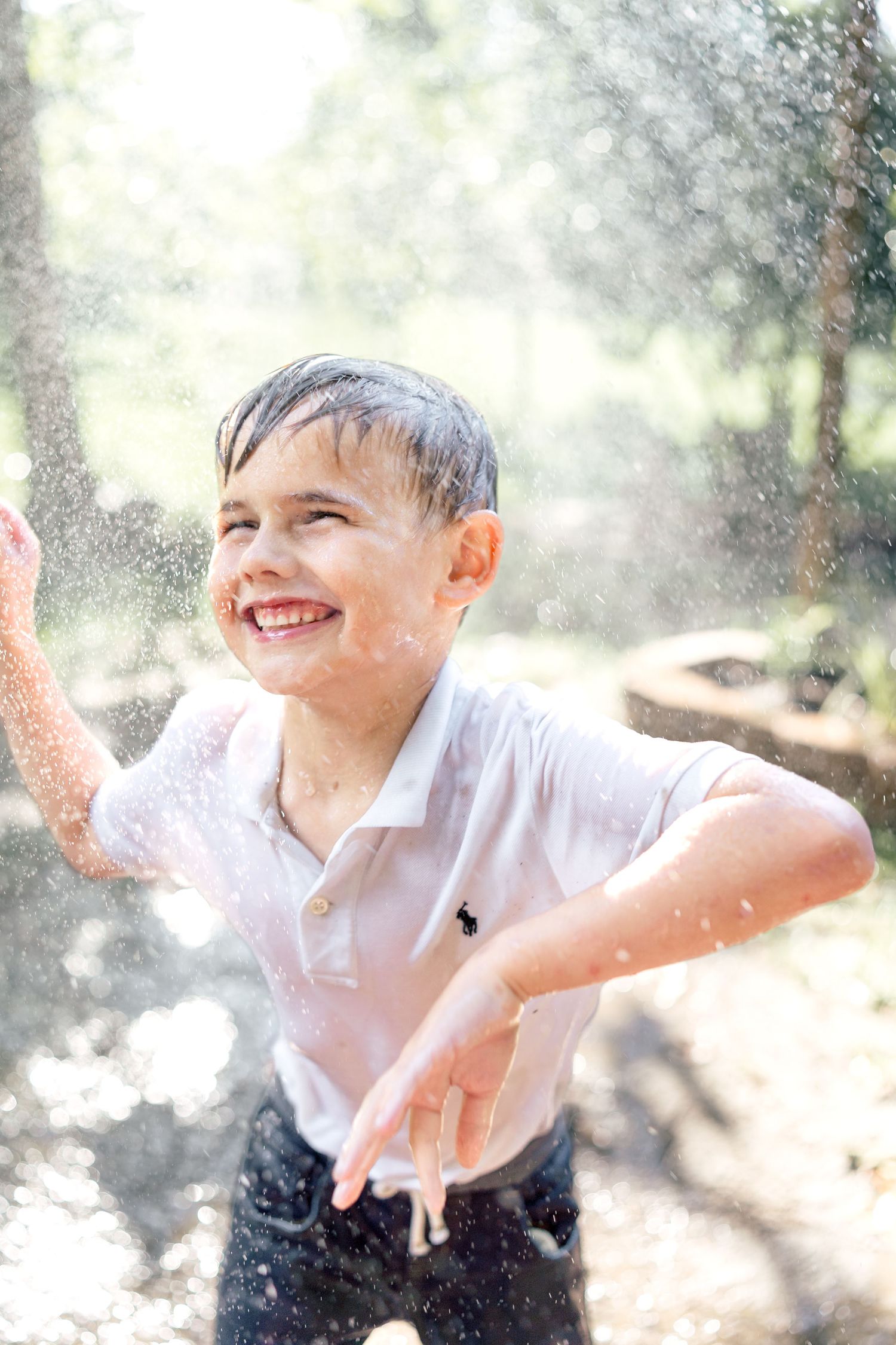 A joyful boy in a white polo shirt laughs while playing in water sprinklers outdoors on a sunny day.