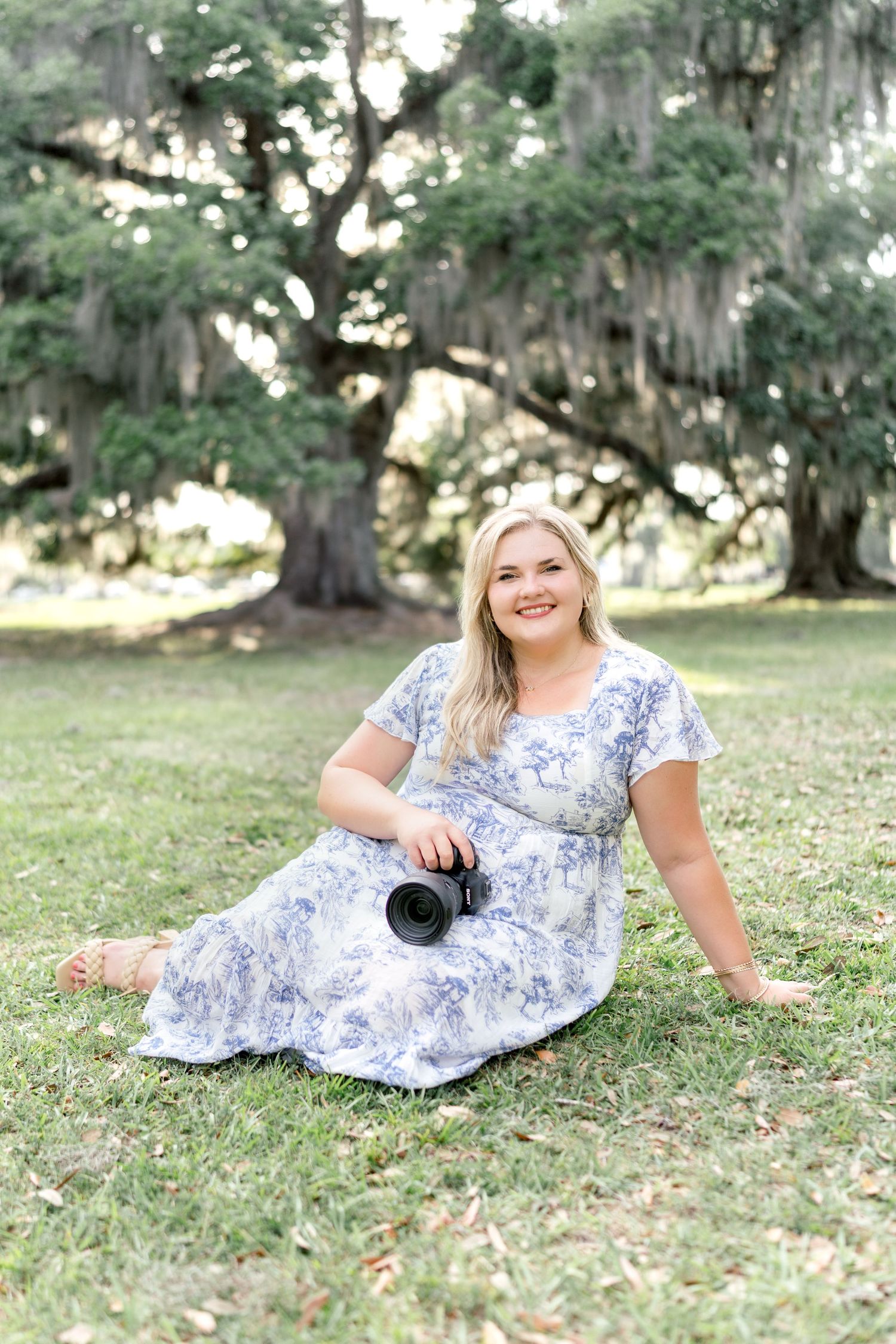 A smiling blonde woman in a blue floral dress sits on grass holding a camera, with large moss-draped oak trees behind her.