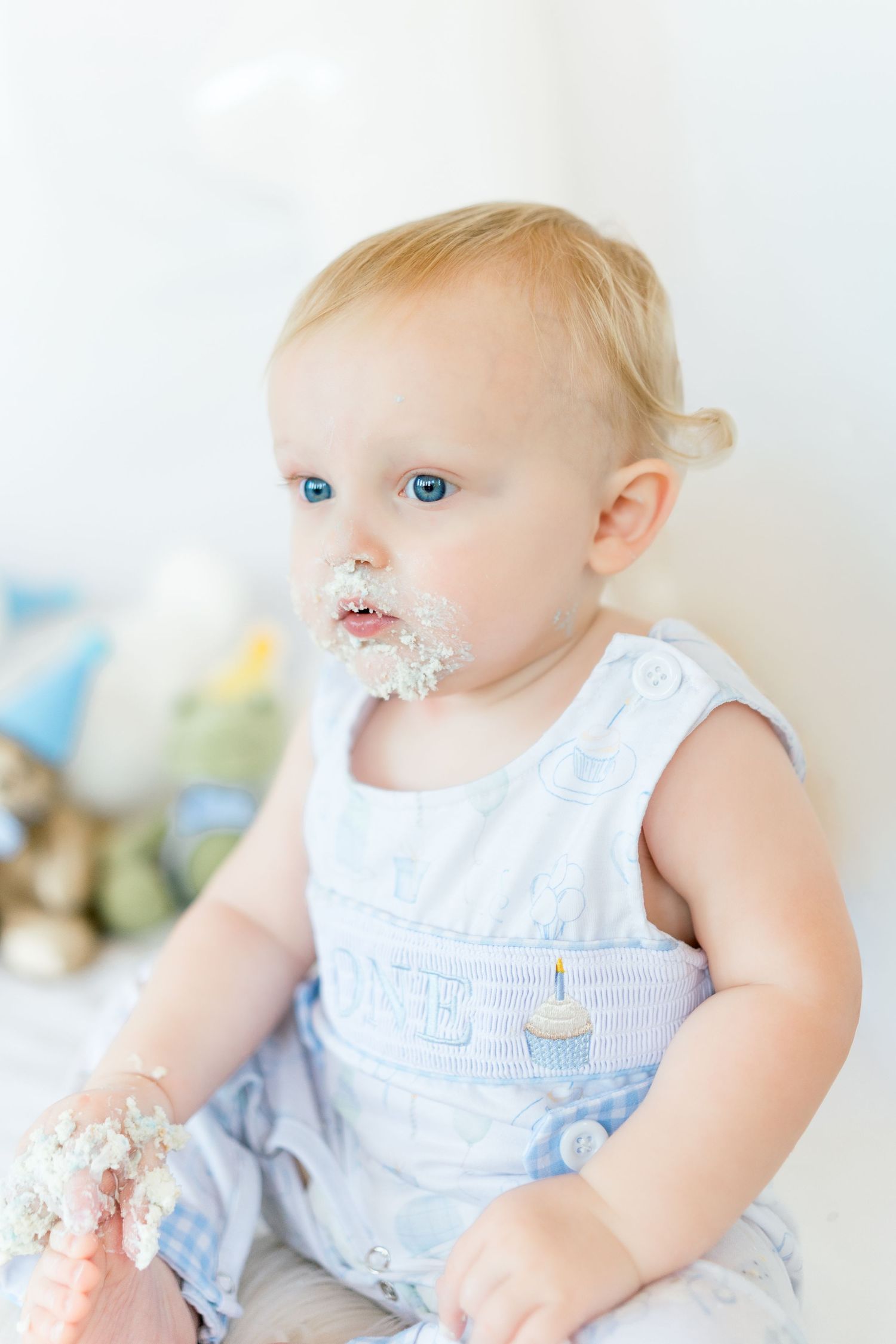 Baby in white sleeveless outfit enjoys messy cake smashing photo session with pink frosting.