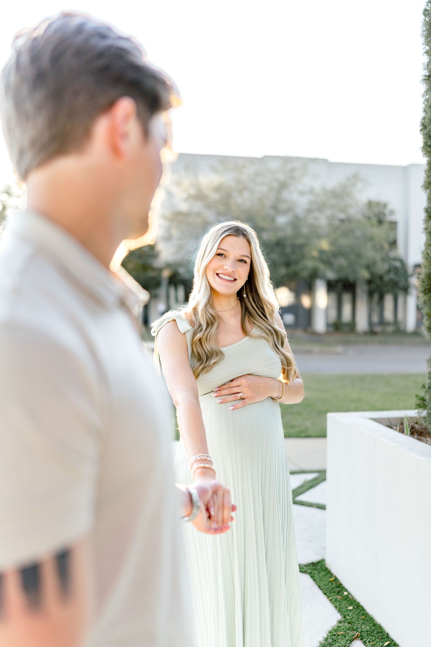 Smiling pregnant woman in mint dress holds belly while reaching for partner's hand outdoors in warm sunlight.