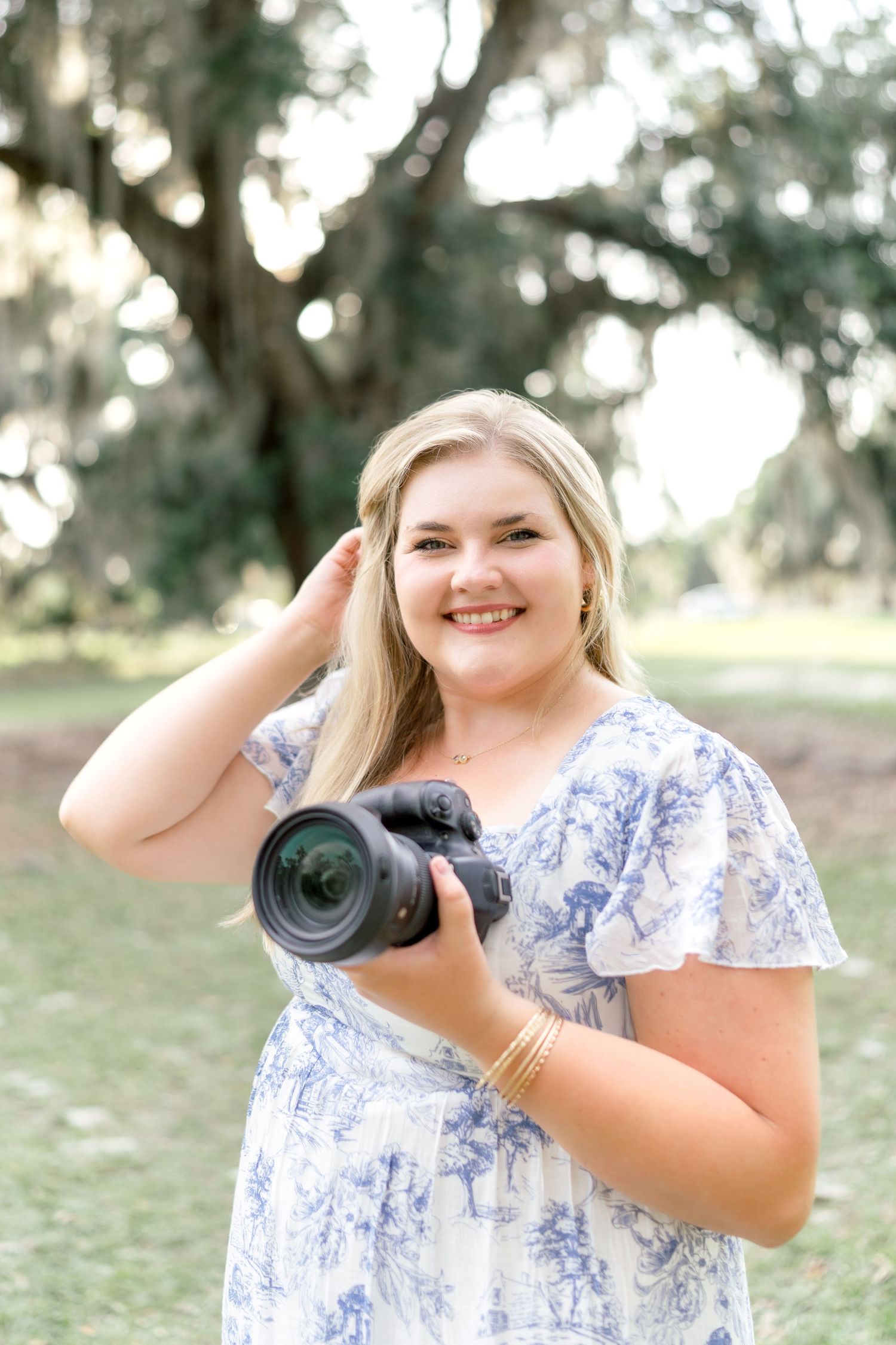 Smiling blonde woman in blue floral dress holding a DSLR camera outdoors under large oak trees.