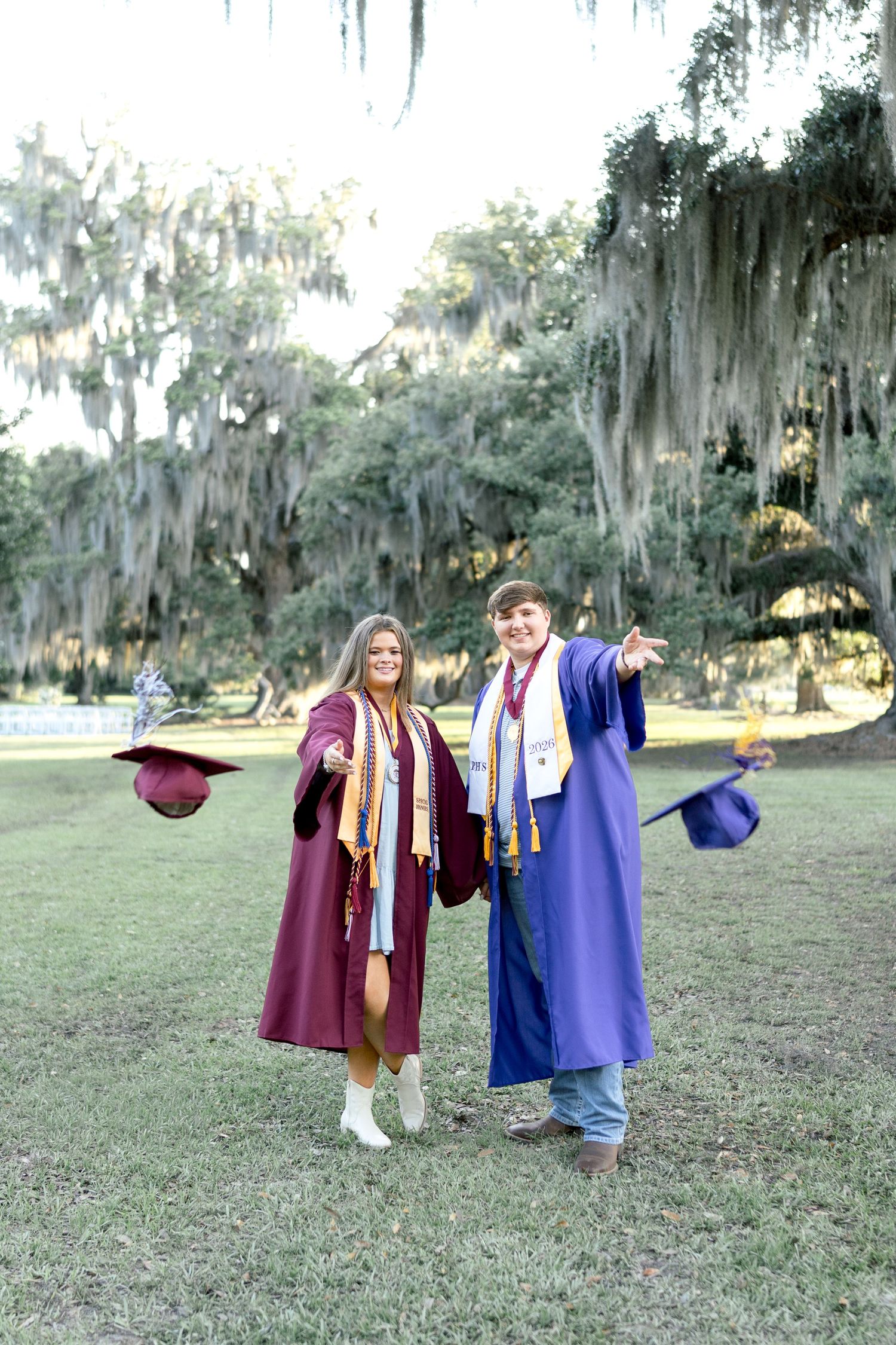 Two high school graduates in maroon and blue caps and gowns celebrate outdoors under Spanish moss-draped trees.