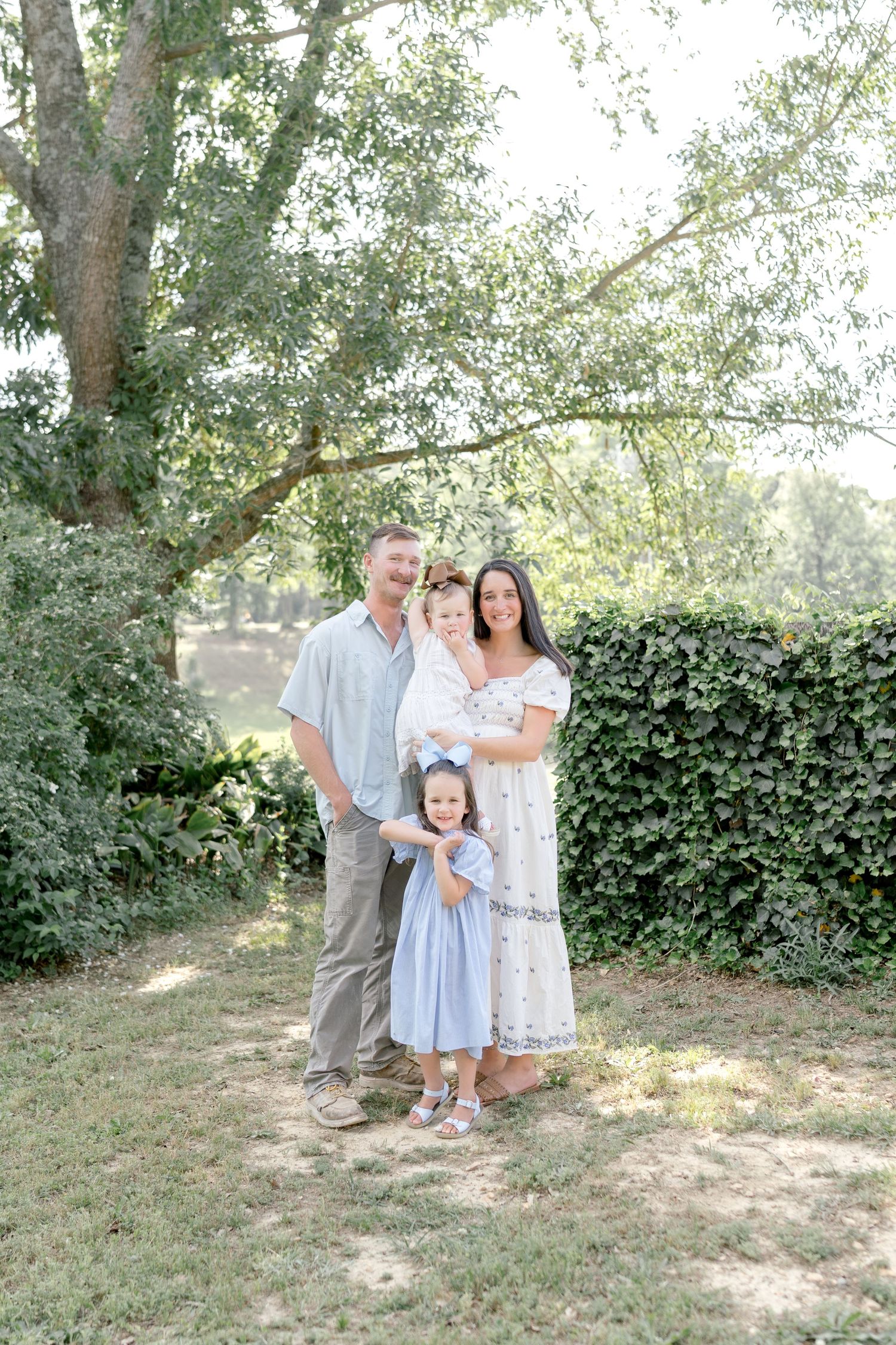 A happy family of four poses outdoors among lush green trees and hedges on a sunny day.